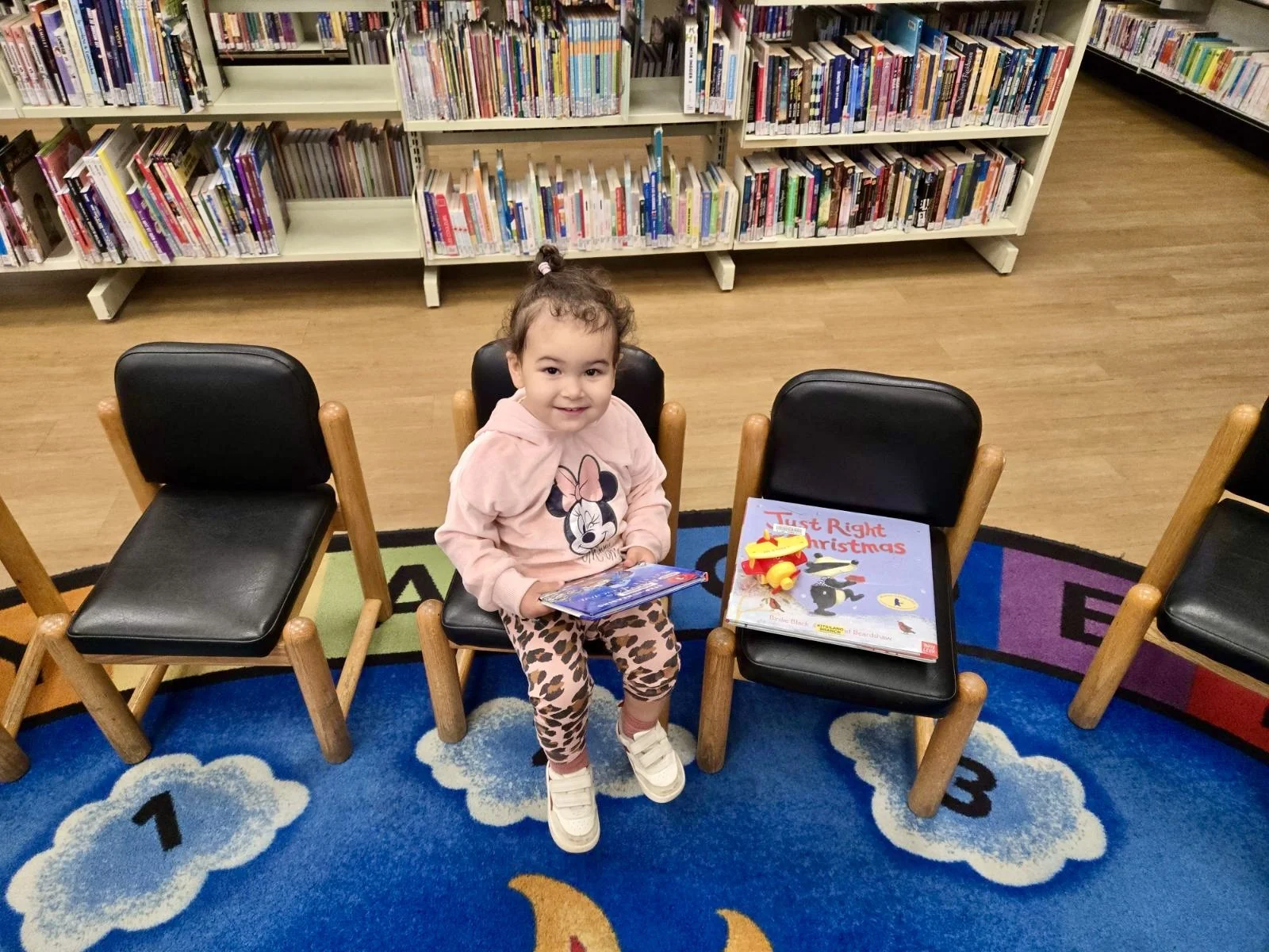ittle girl dressed in mini mouse seater sitting on a small chair while looking at books in the library