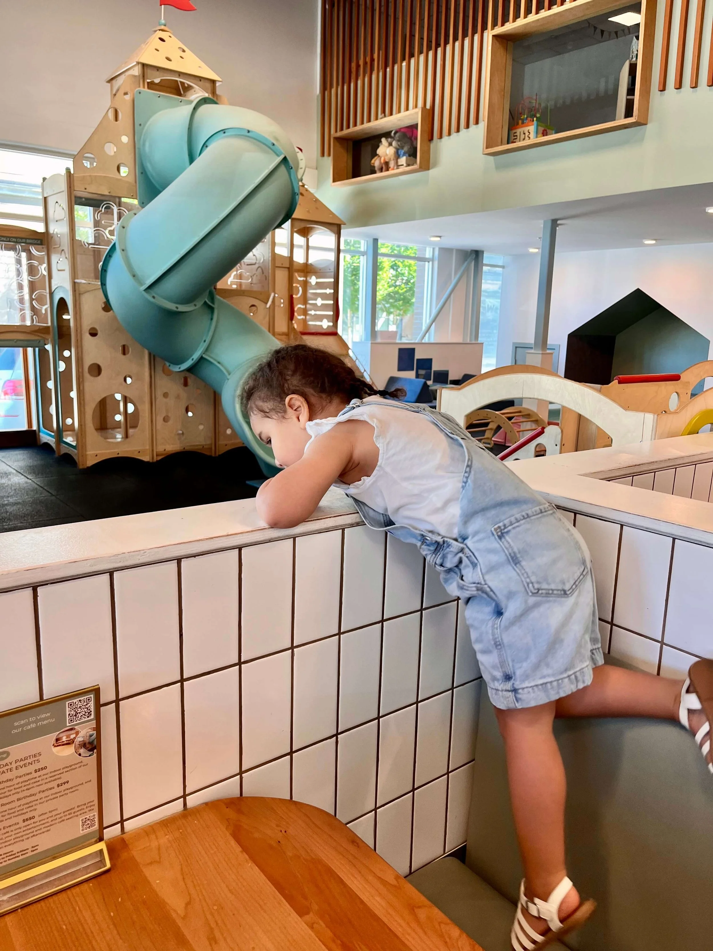 Little girl standing in a cafe booth and leaning over the side to look at the big play area on the other side.