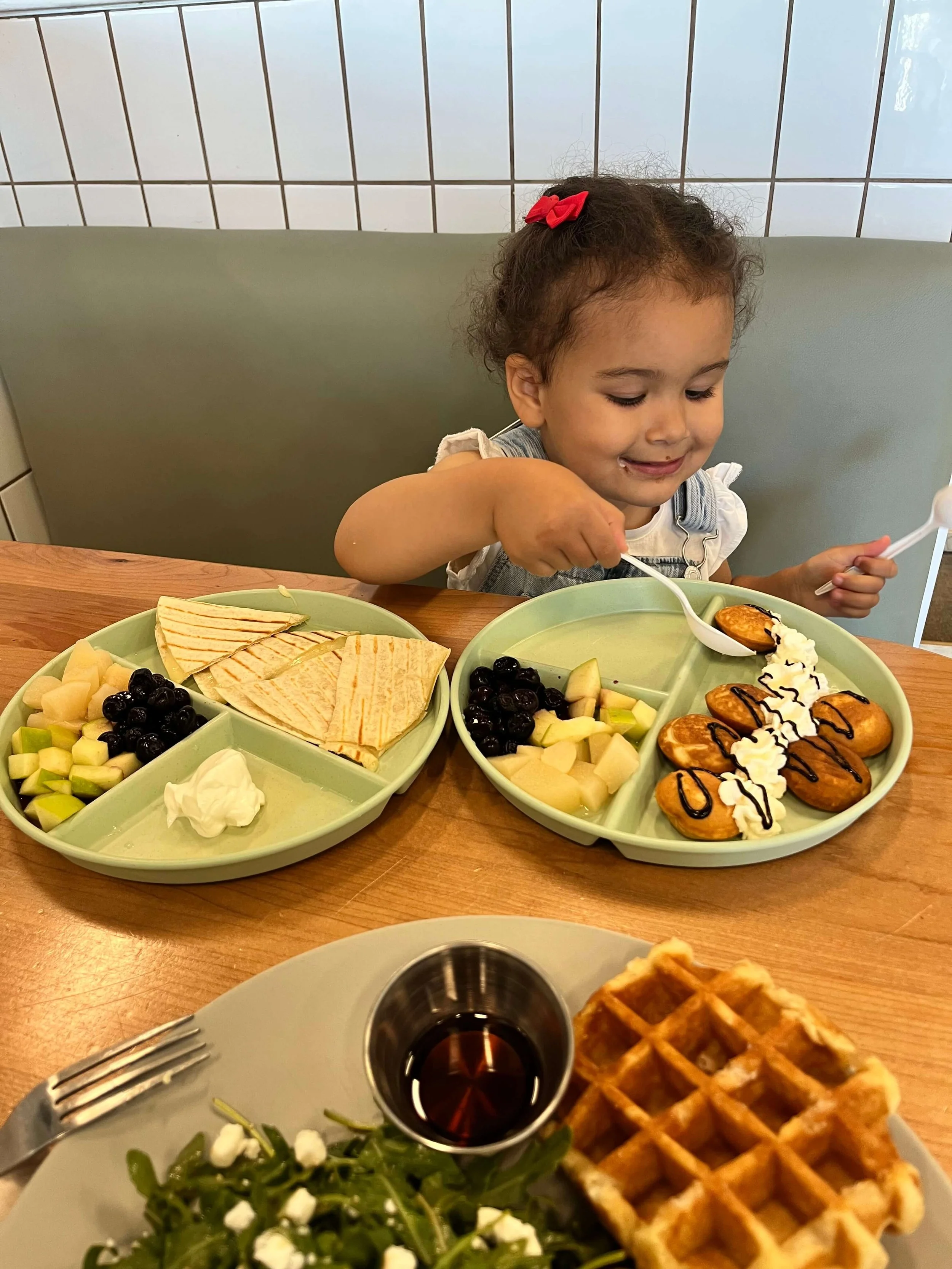 little girl dressed in overalls sitting at a cafe table surrounded by two plates filled with food.