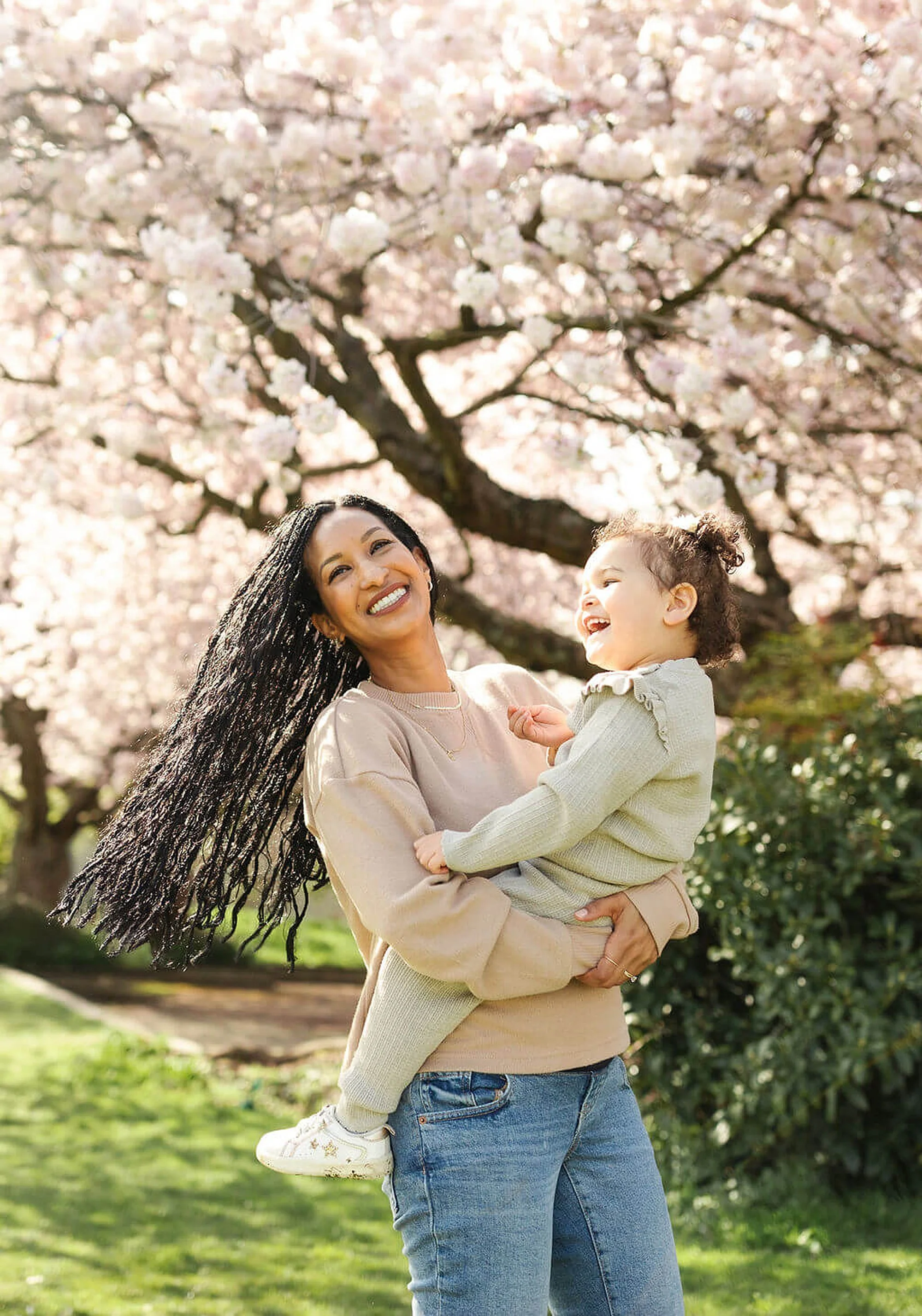A woman holding a young girl outdoors with a blooming cherry blossom tree in the background. The woman has long black curly hair and is wearing a beige sweater, while the girl has curly hair and is wearing a light green sweater and light-colored sneakers. They are both smiling and enjoying a sunny day in a park or garden.