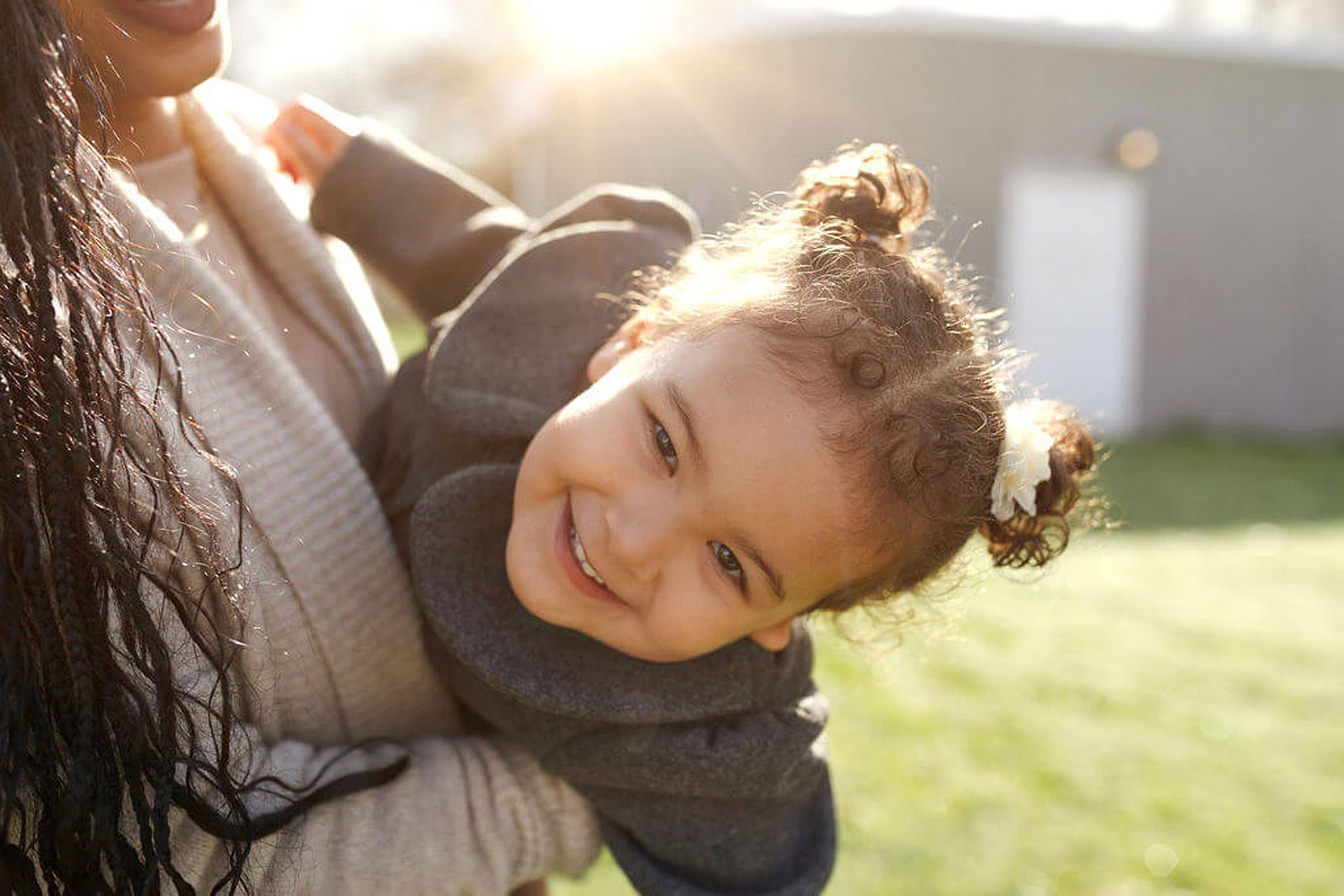 A woman with long, dark, curly hair holding a young girl with curly hair tied in a bun, both smiling outdoors in sunlight.