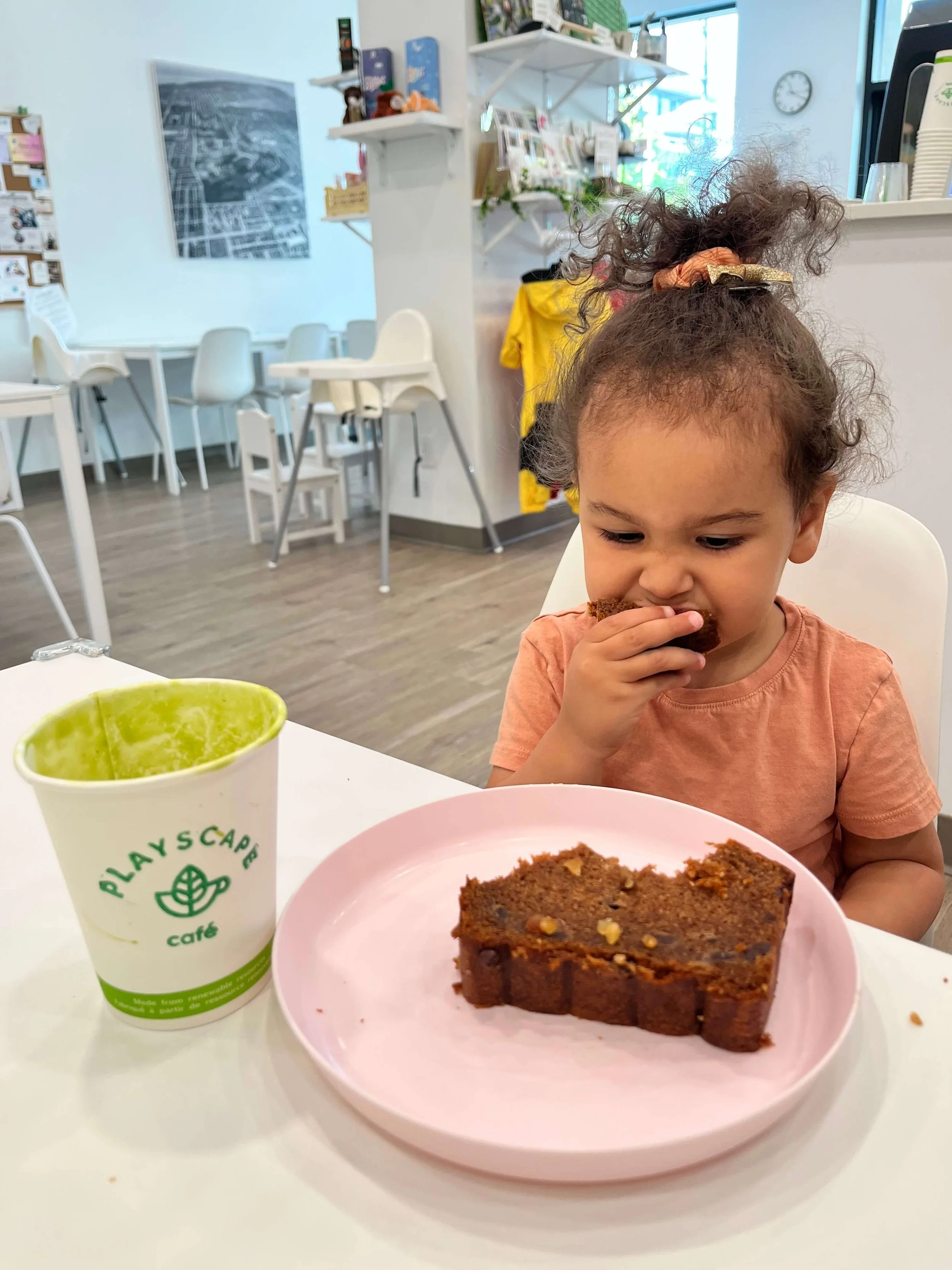 little girl in pink shirt sitting at a table. She is eating a banana bread slice and a green smooties is next to her.
