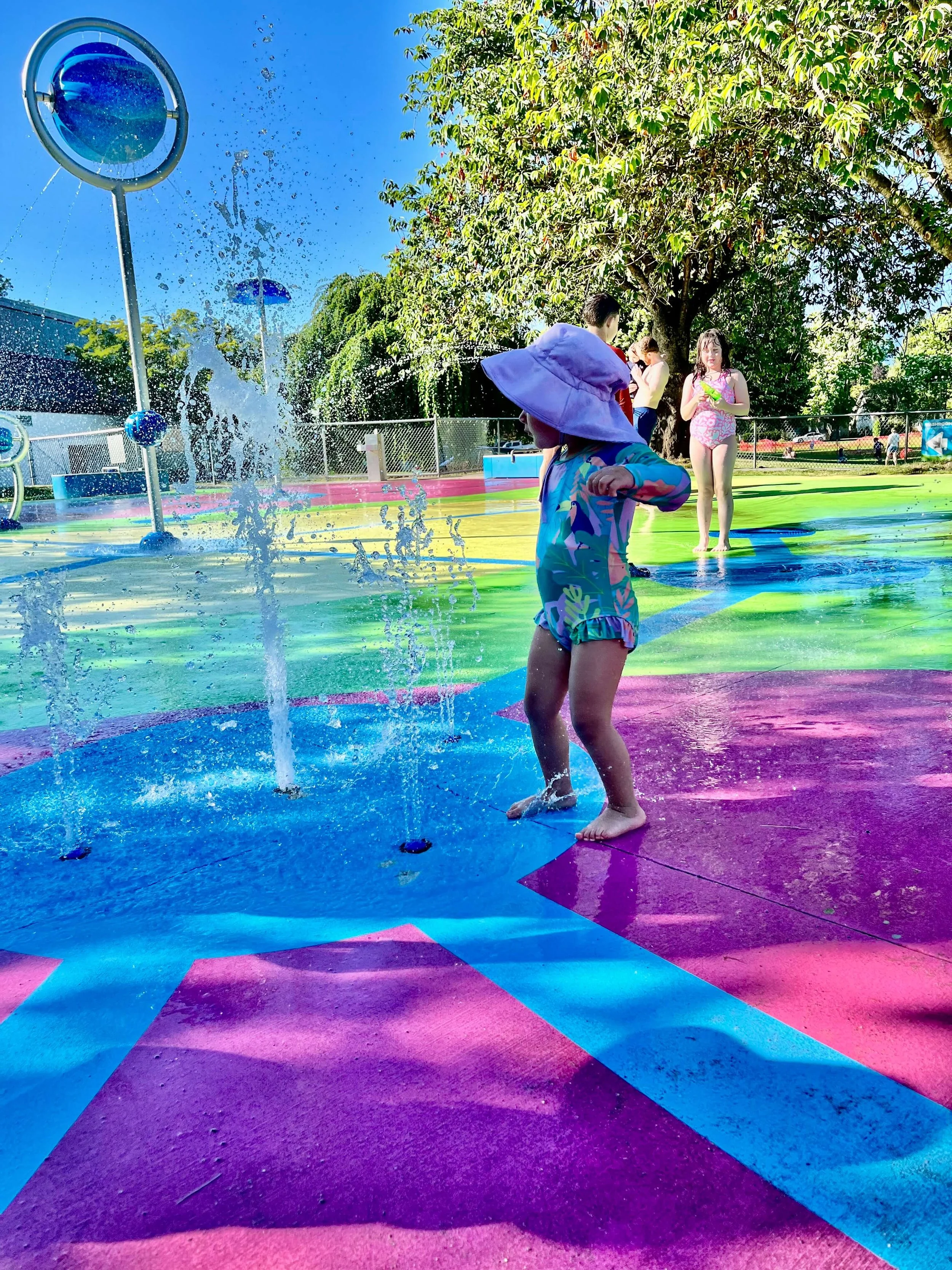 little girl in a purple hat and tropical print bathing suit running in the water park.