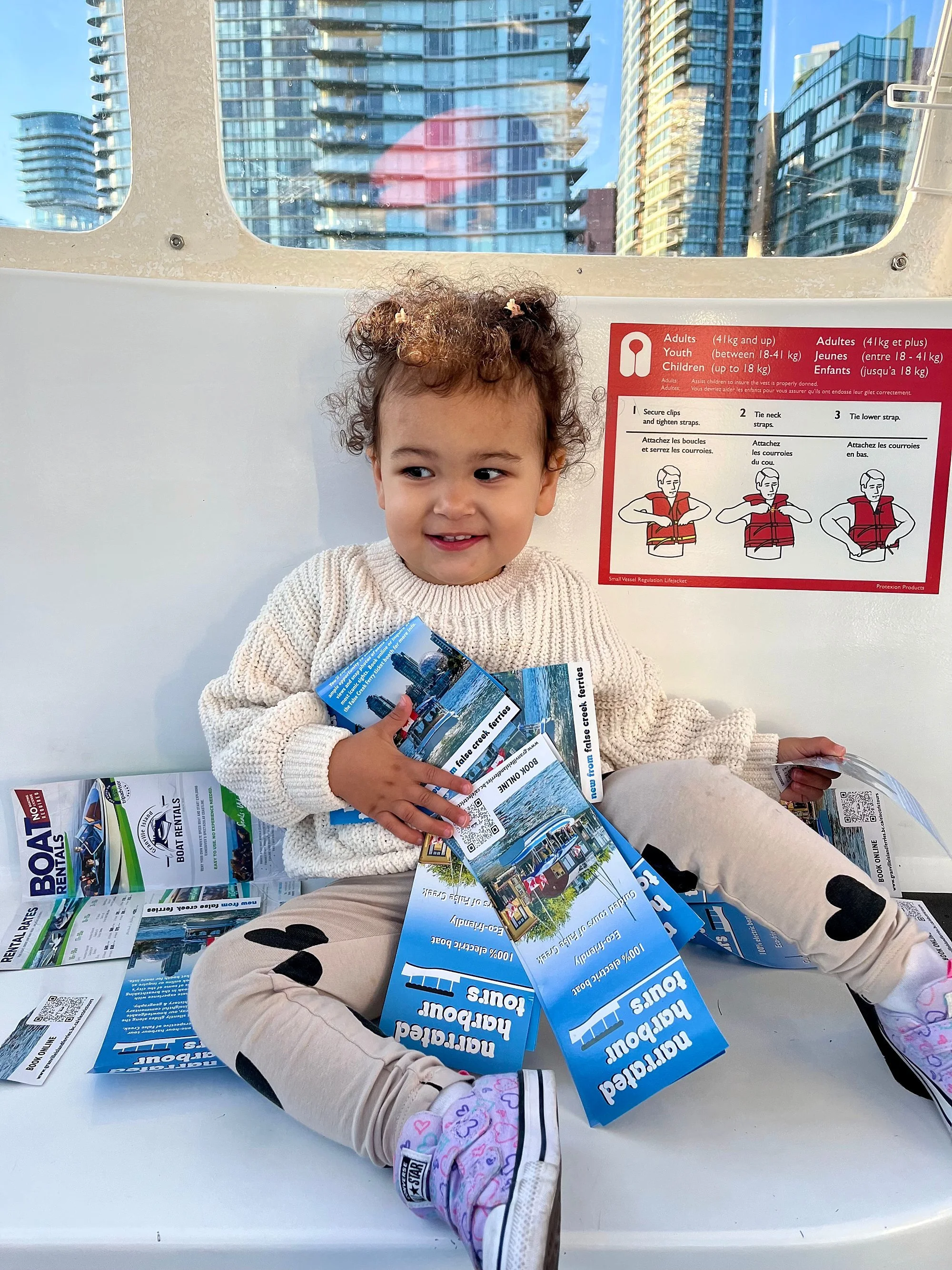 A young girl sitting on the floor with a large number of brochures and pamphlets on her lap, surrounded by more brochures, in an urban setting with tall buildings visible through a window behind her.