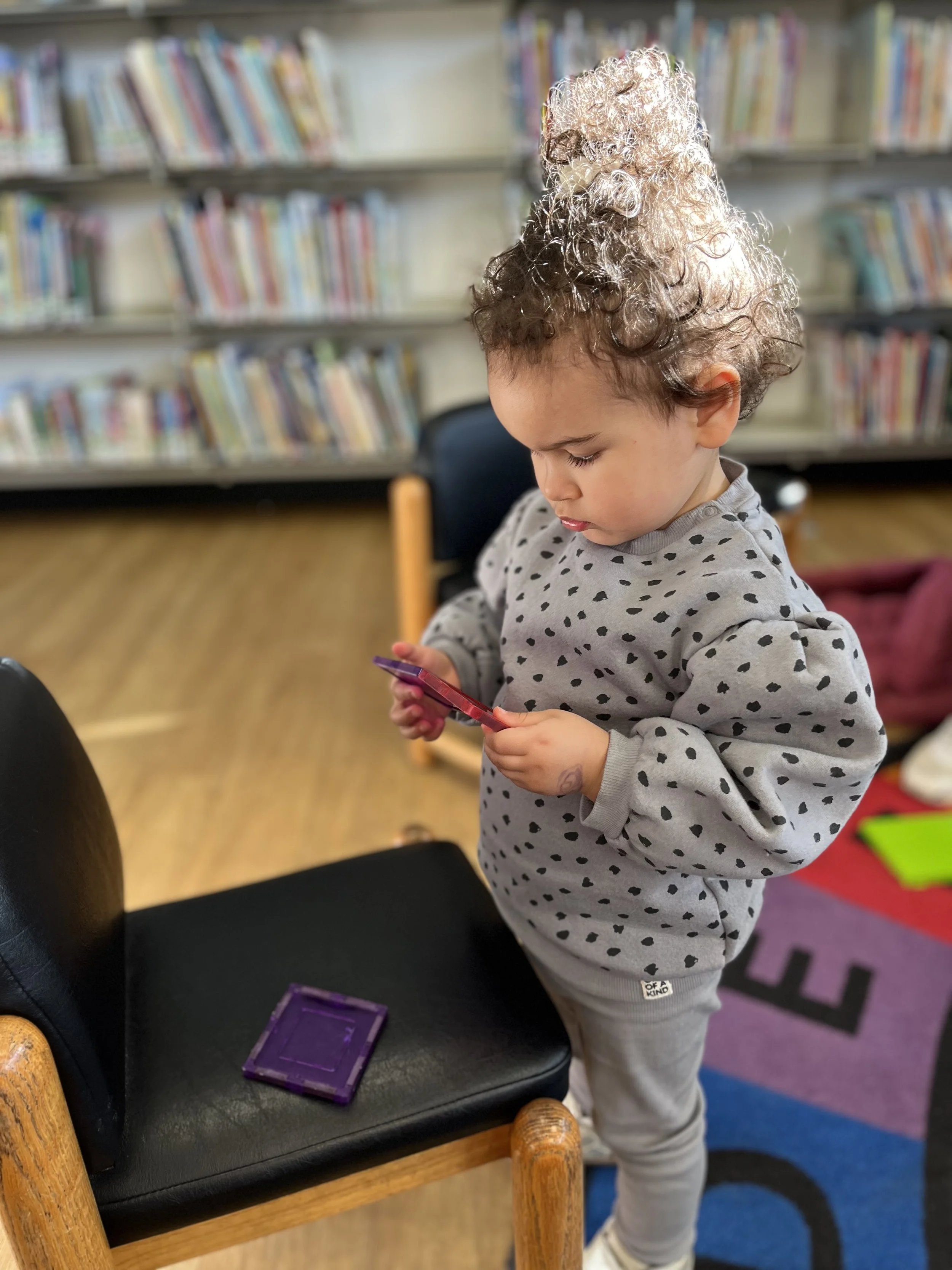 little girl in a grey sweater concentrating and playing with magnetic tiles.