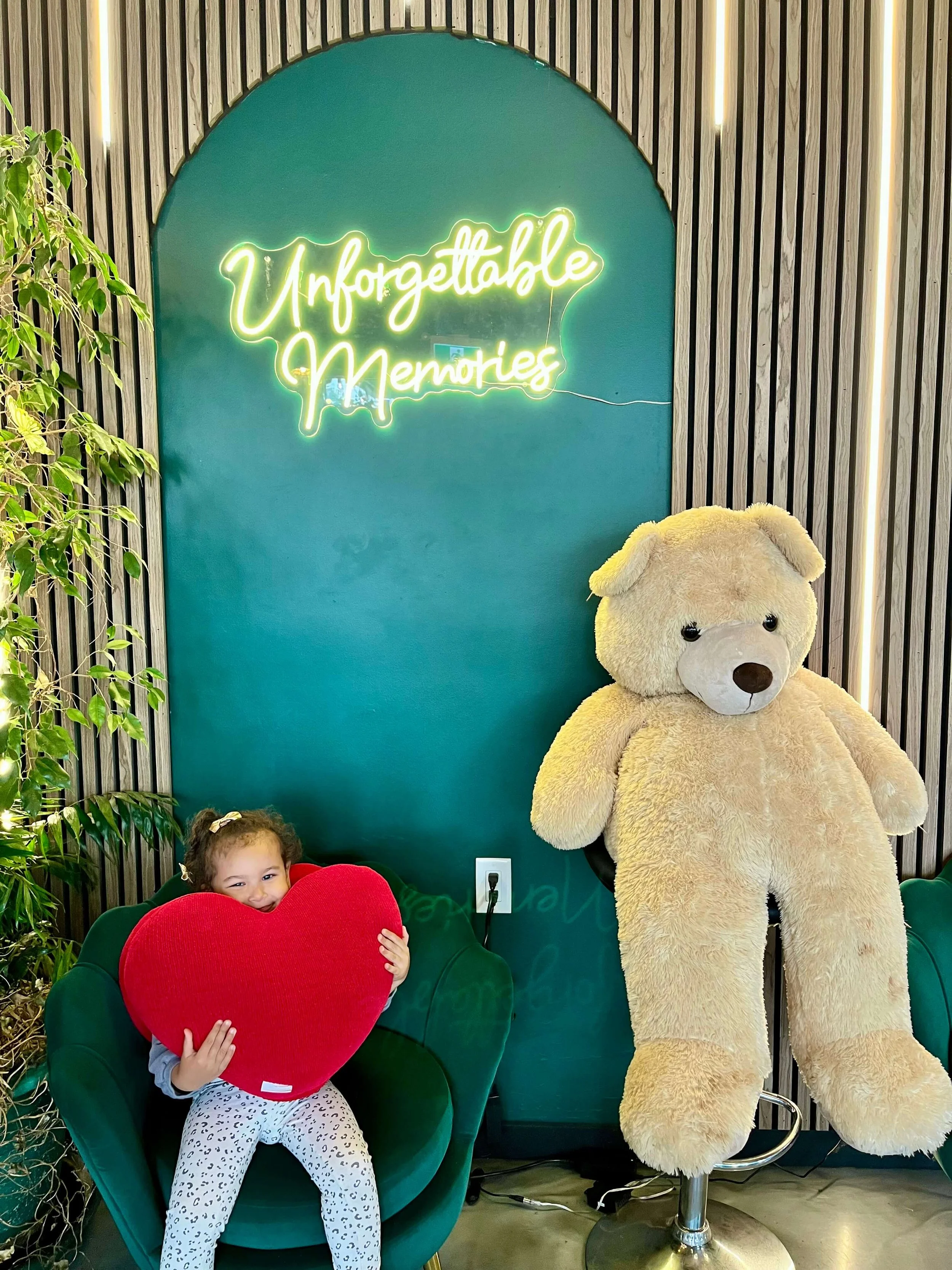 Little girl sitting on a green chair holding a big read heart cushion next to a big teddy bear