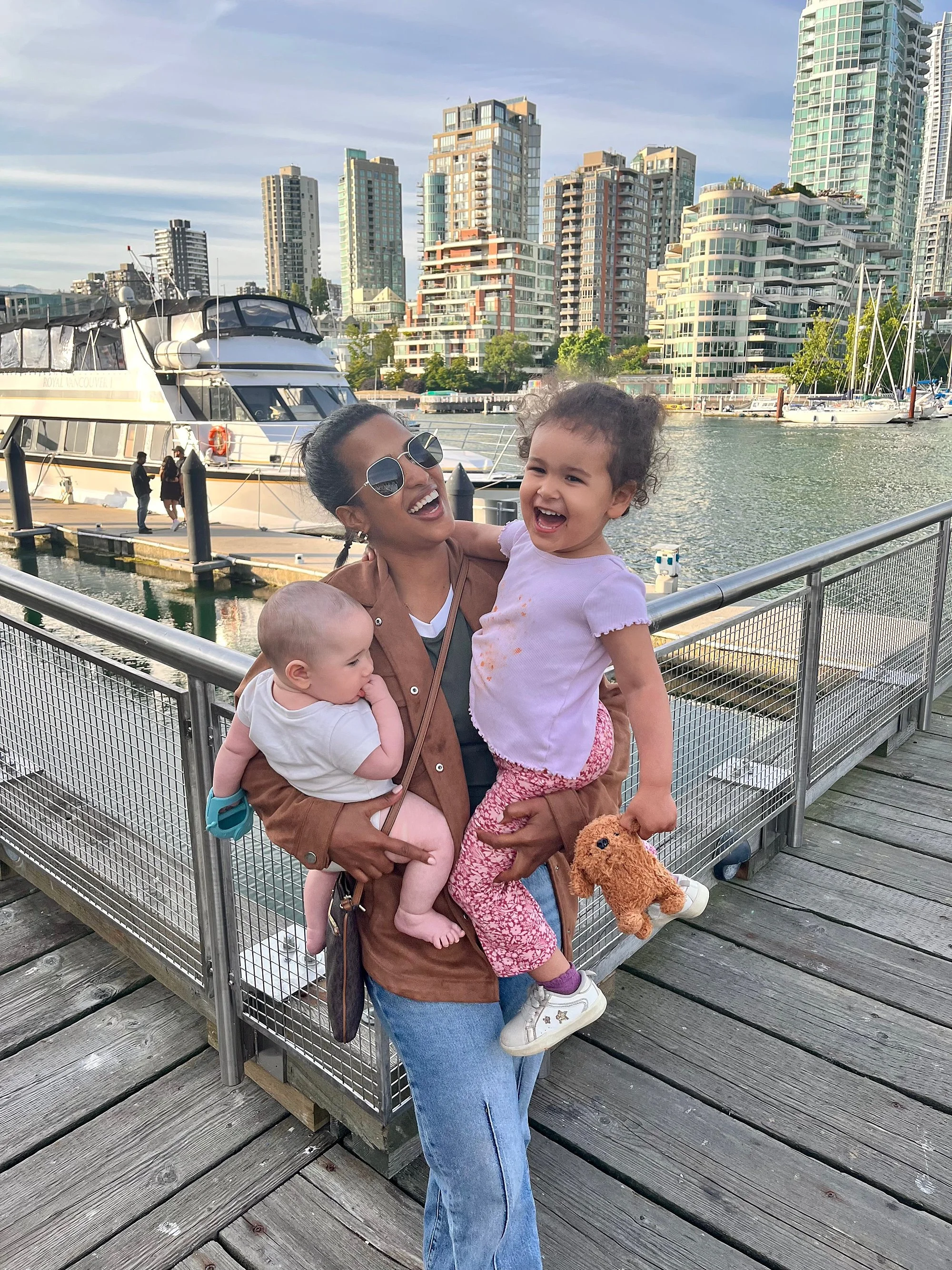 A woman with sunglasses holding two young children on a wooden pier near the water, with boats and modern high-rise buildings in the background.