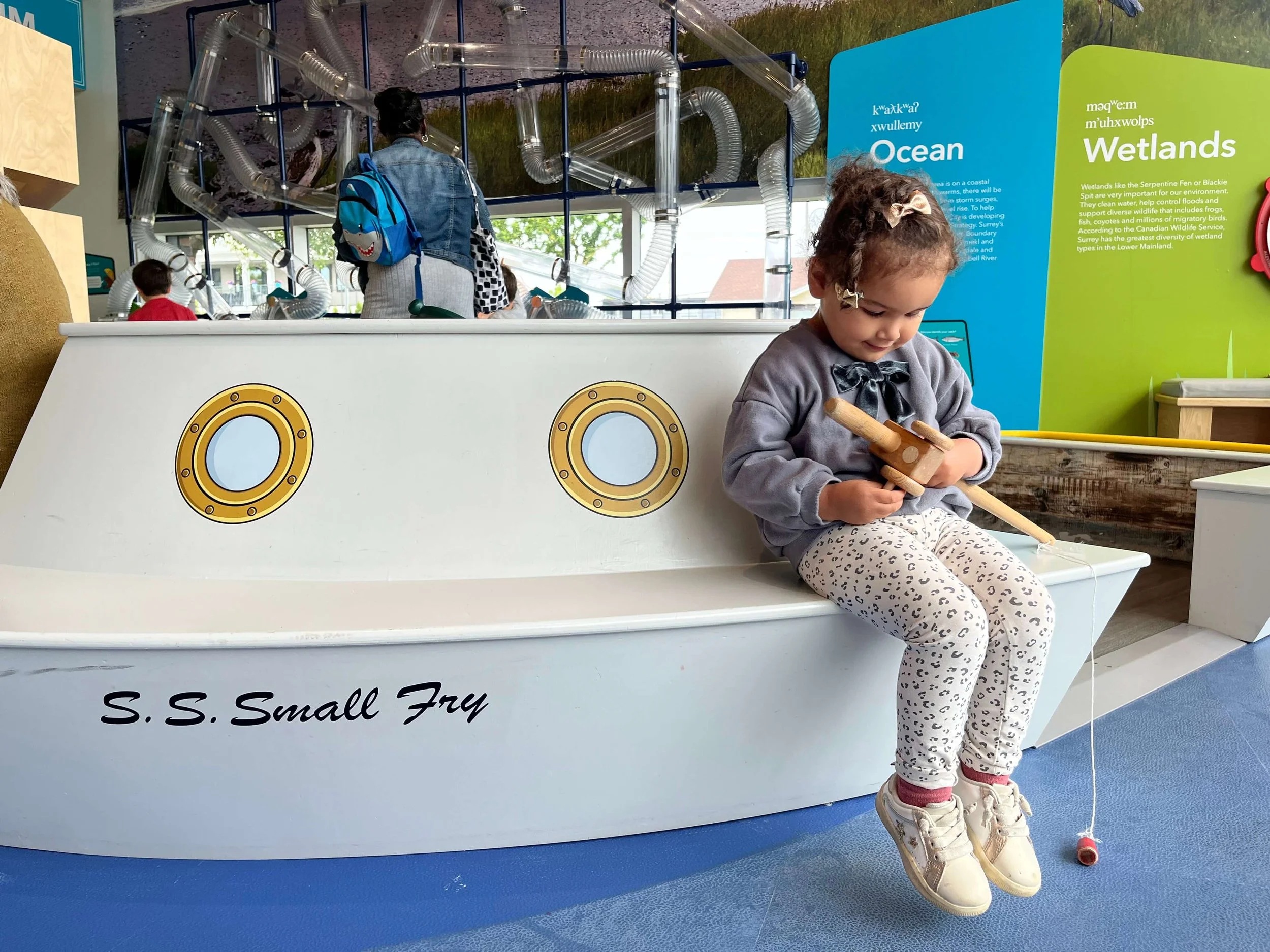 Little girl sitting on the edge of a toy fishing boat trying to learn how to use the fishing rod