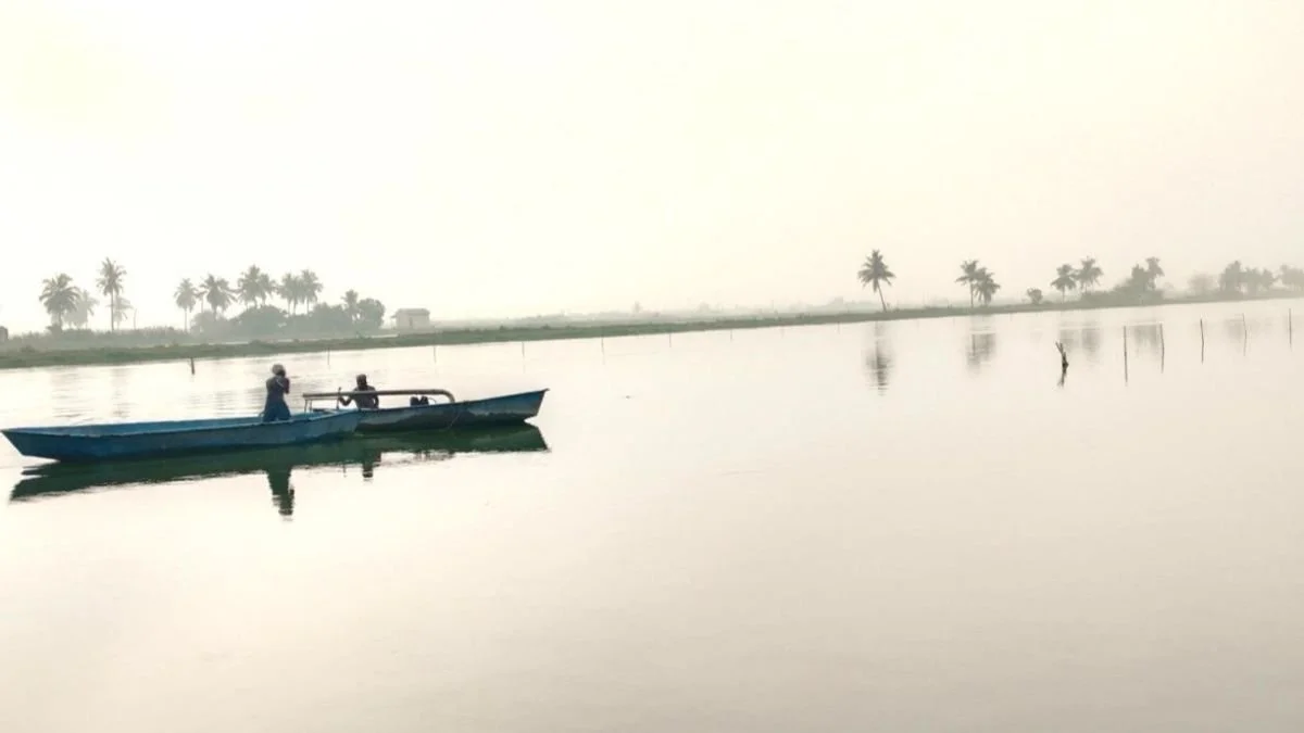 A boat with two people on a calm river with palm trees in the background.