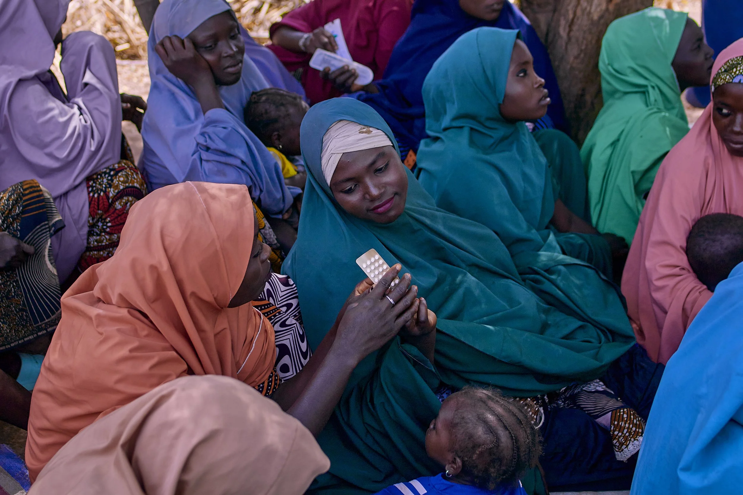 A group of women sitting outdoors, some wearing colorful hijabs and traditional clothing, with one woman in the foreground holding a blister pack of pills.
