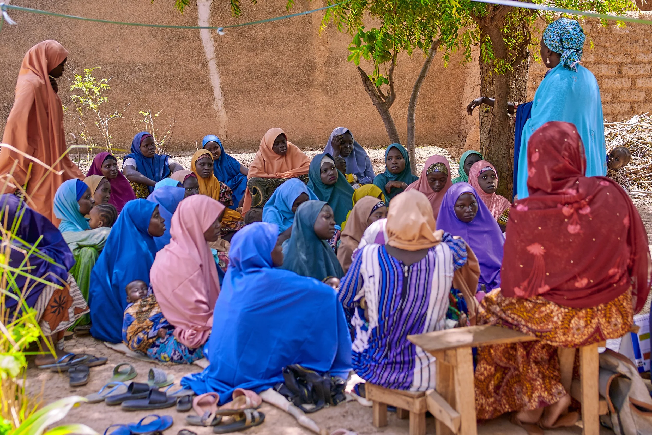 Group of women and children sitting outdoors, listening to a woman in a blue dress who appears to be speaking. The women are dressed in colorful traditional clothing with headscarves, and shoes are placed on the ground nearby. A tree provides shade, and a brick wall is in the background.
