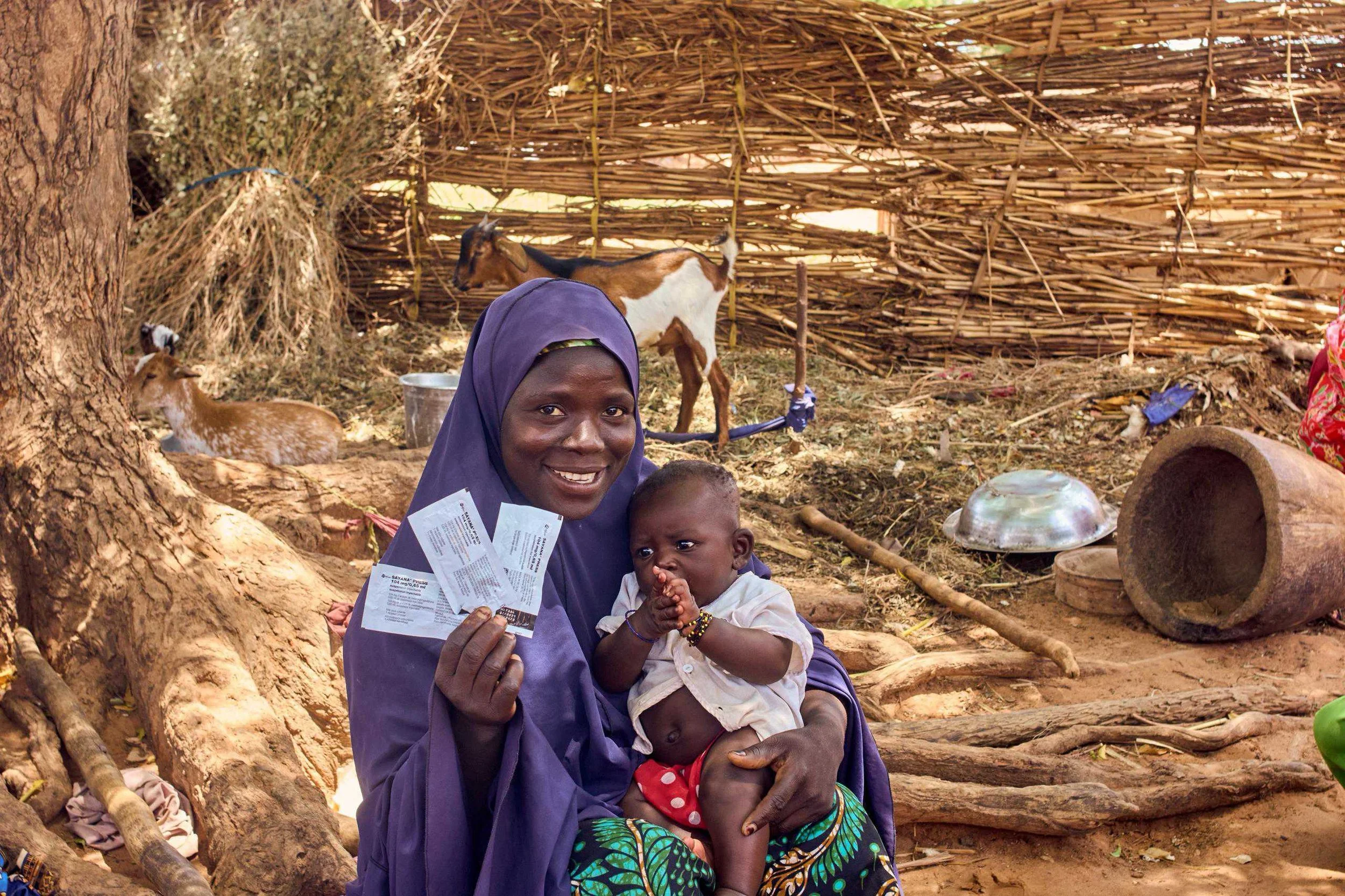 A smiling woman in a purple headscarf holds two children, sitting outdoors in front of a woven fence with goats and farm supplies around them.