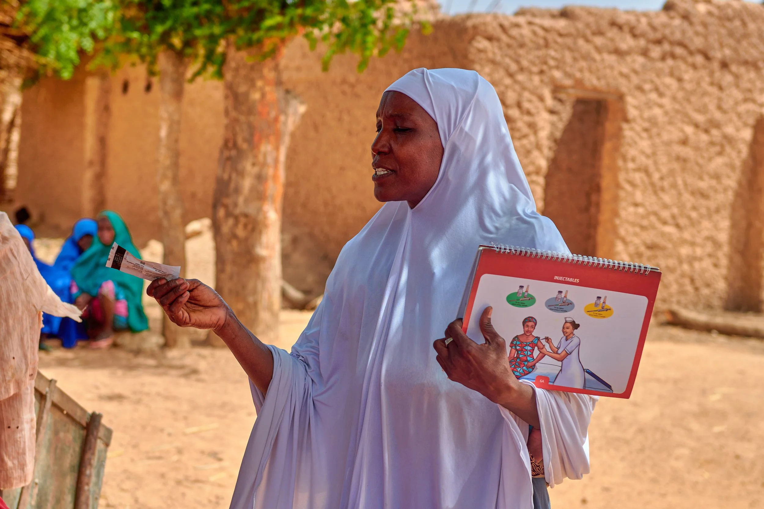 A woman dressed in white, wearing a headscarf, holds a medical pamphlet and a clipboard with medical illustrations while speaking to a person outside, with a dirt wall and other women in colorful clothing in the background.
