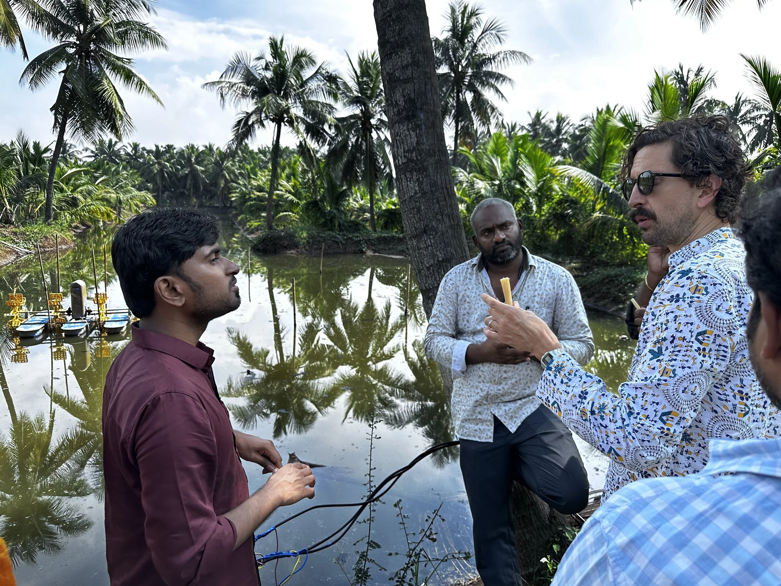Three men engaged in a conversation outdoors near a water body, surrounded by lush green coconut trees, with one of them holding a fishing rod.