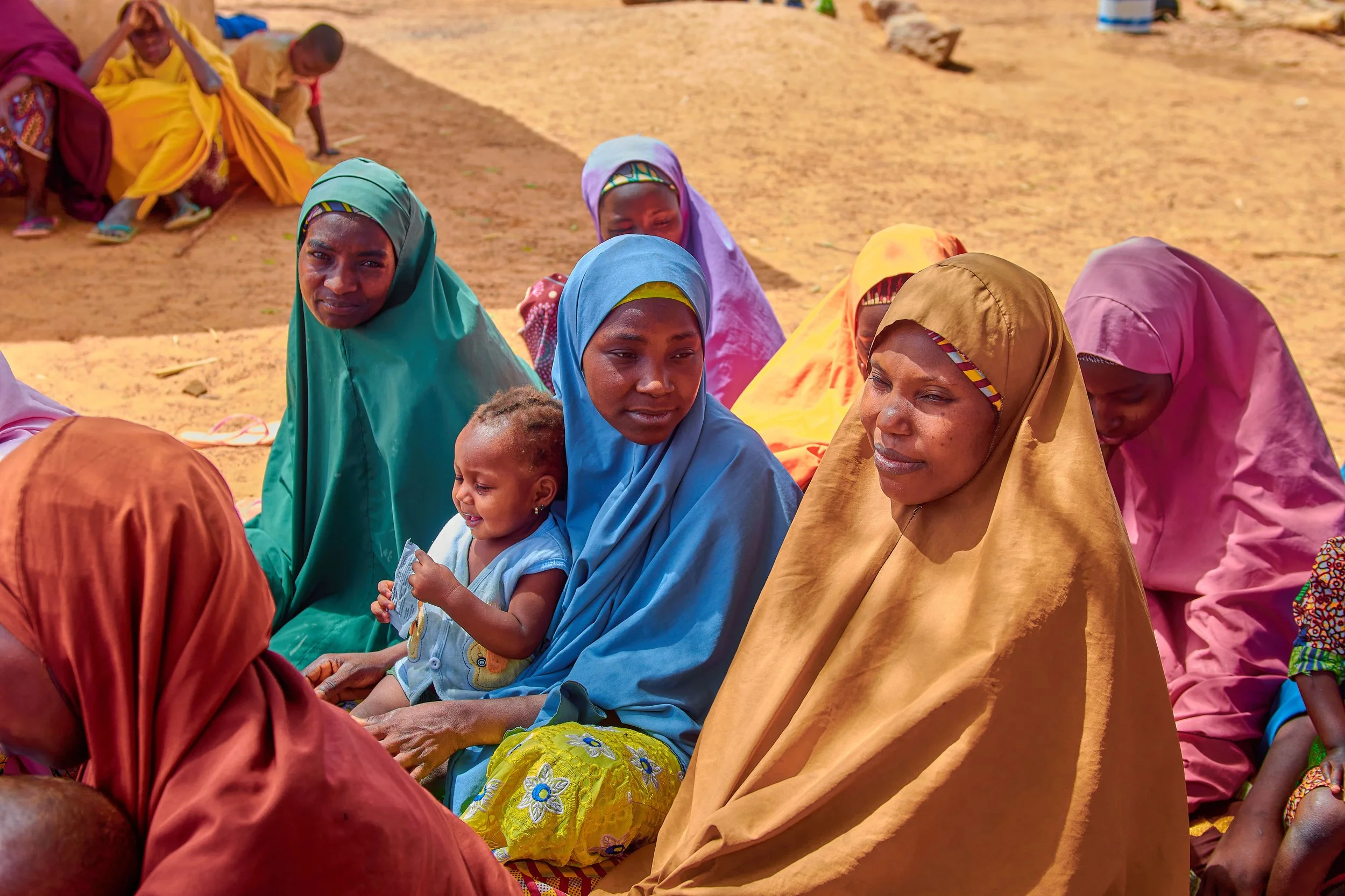 Group of women and children sitting outdoors on dirt ground, wearing colorful traditional clothing and head coverings, with some looking at the camera and others looking downward.