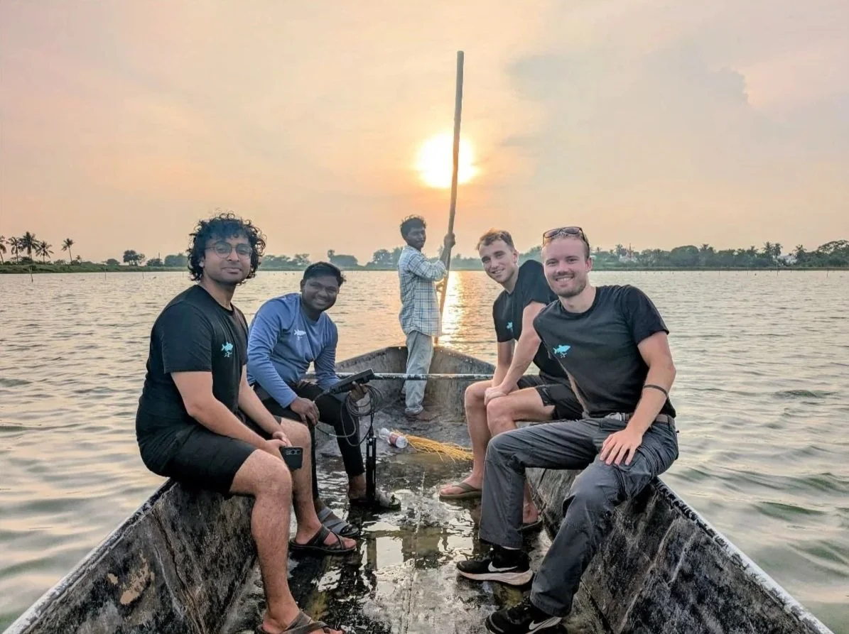 Group of five men on a boat during sunset on a water body with a distant shoreline of trees and palm trees.