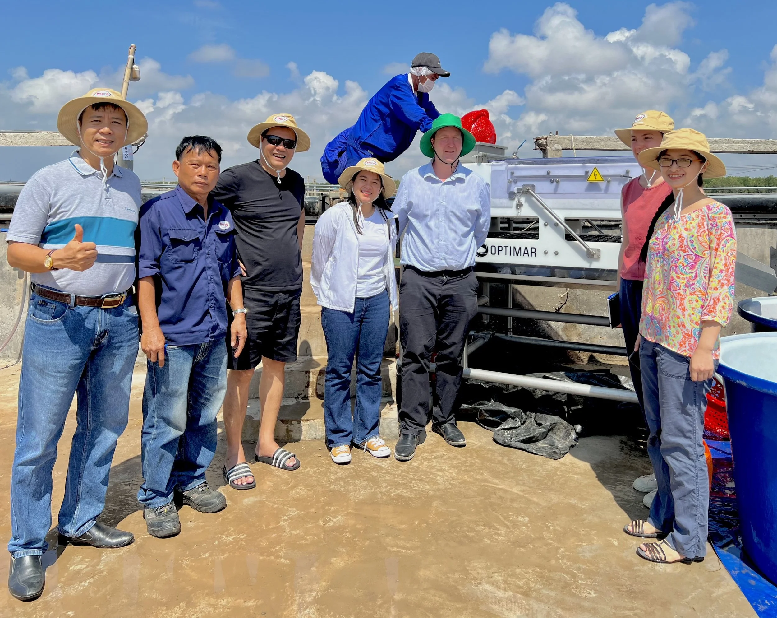 Group of people standing outdoors on a sunny day, wearing hats and masks, near equipment with an industrial or scientific setting in the background.