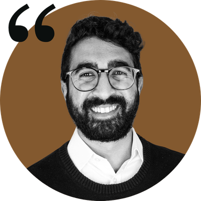 A man with glasses and a beard smiling, wearing a white shirt and dark sweater, against a brown background.