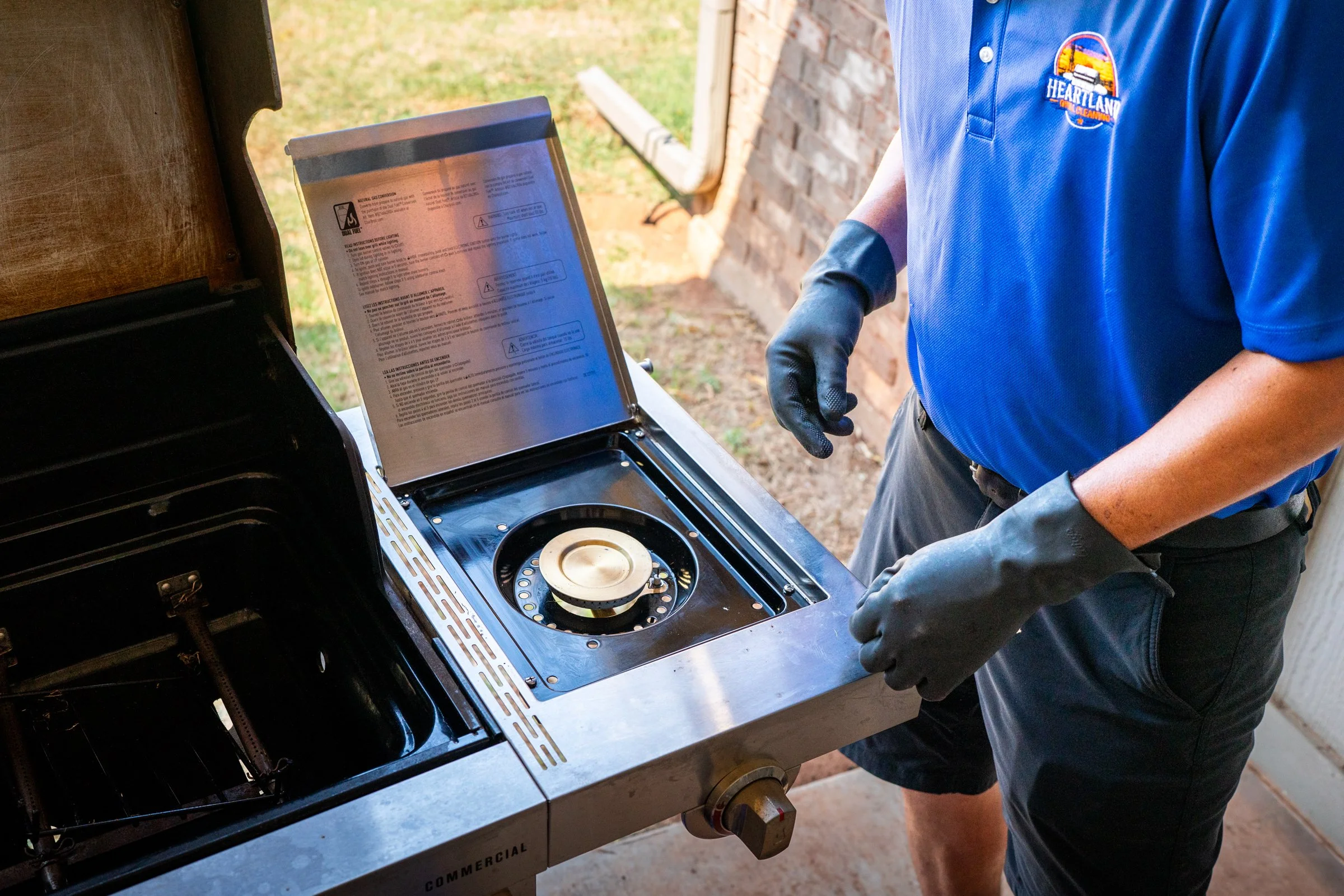 Person wearing a blue Heartland logo shirt and black gloves adjusts a black gas grill in an outdoor setting with a backyard patio in the background.