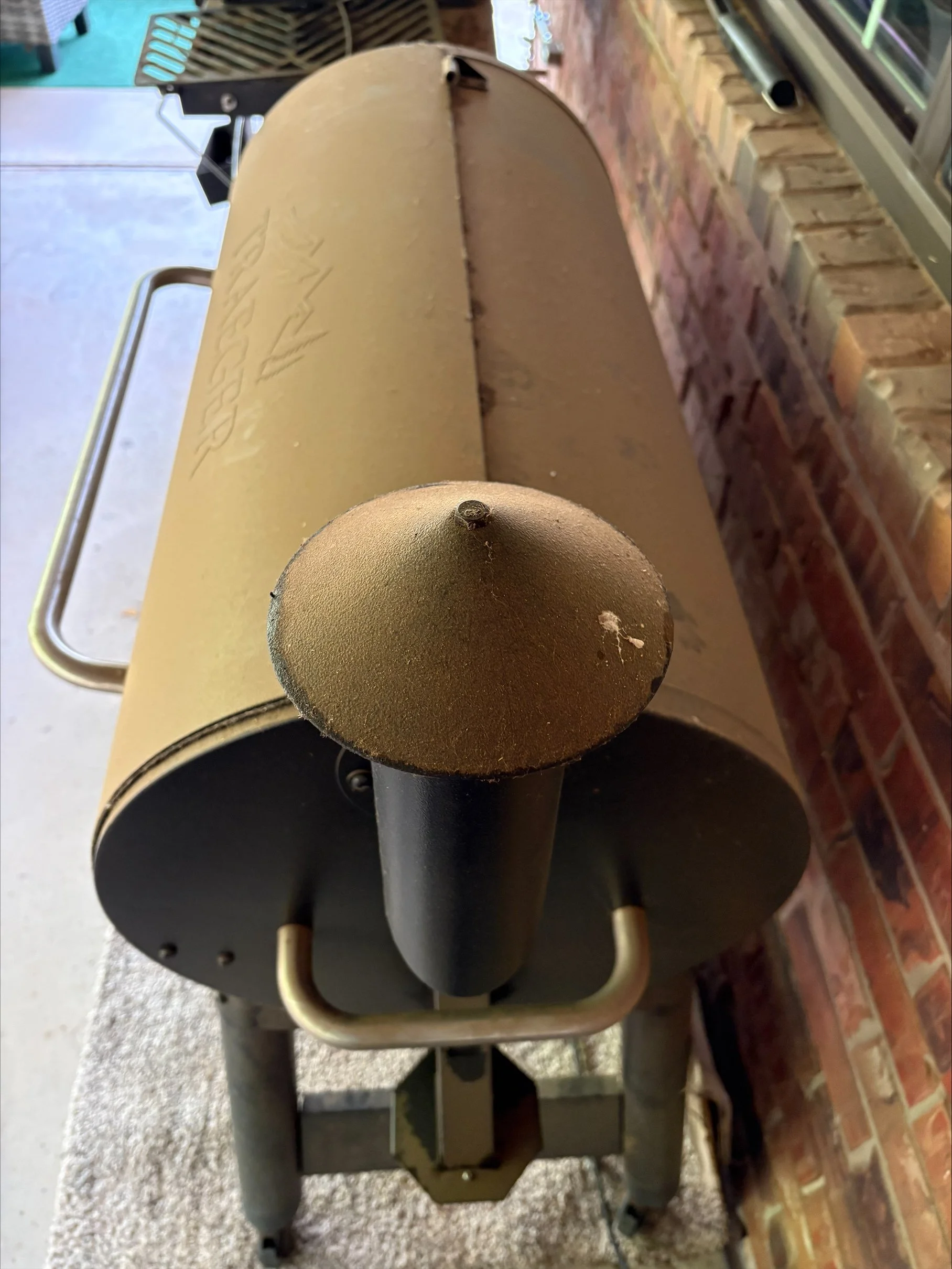 A vintage brick outdoor grill with a rounded vent pipe on top showing signs of dust and caked-on grime as well as cobwebs, situated against a brick wall and adjacent to a freezer or refrigerator.