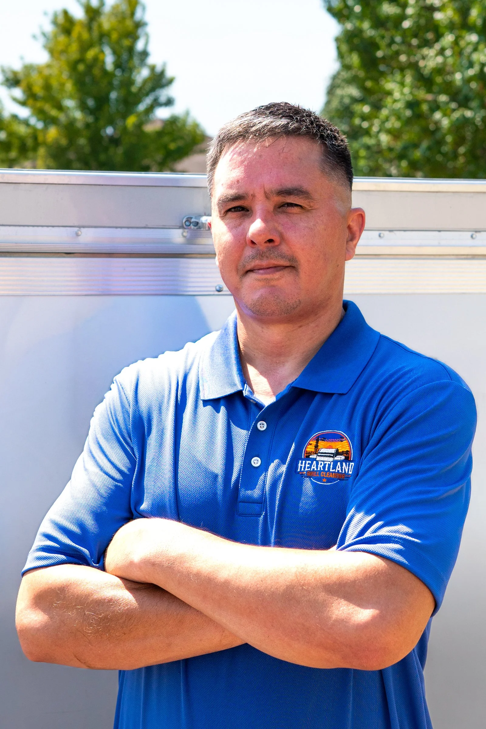 A man wearing a blue polo shirt with a Heartland Grill & Catering logo, arms crossed, standing outdoors with green trees in the background.