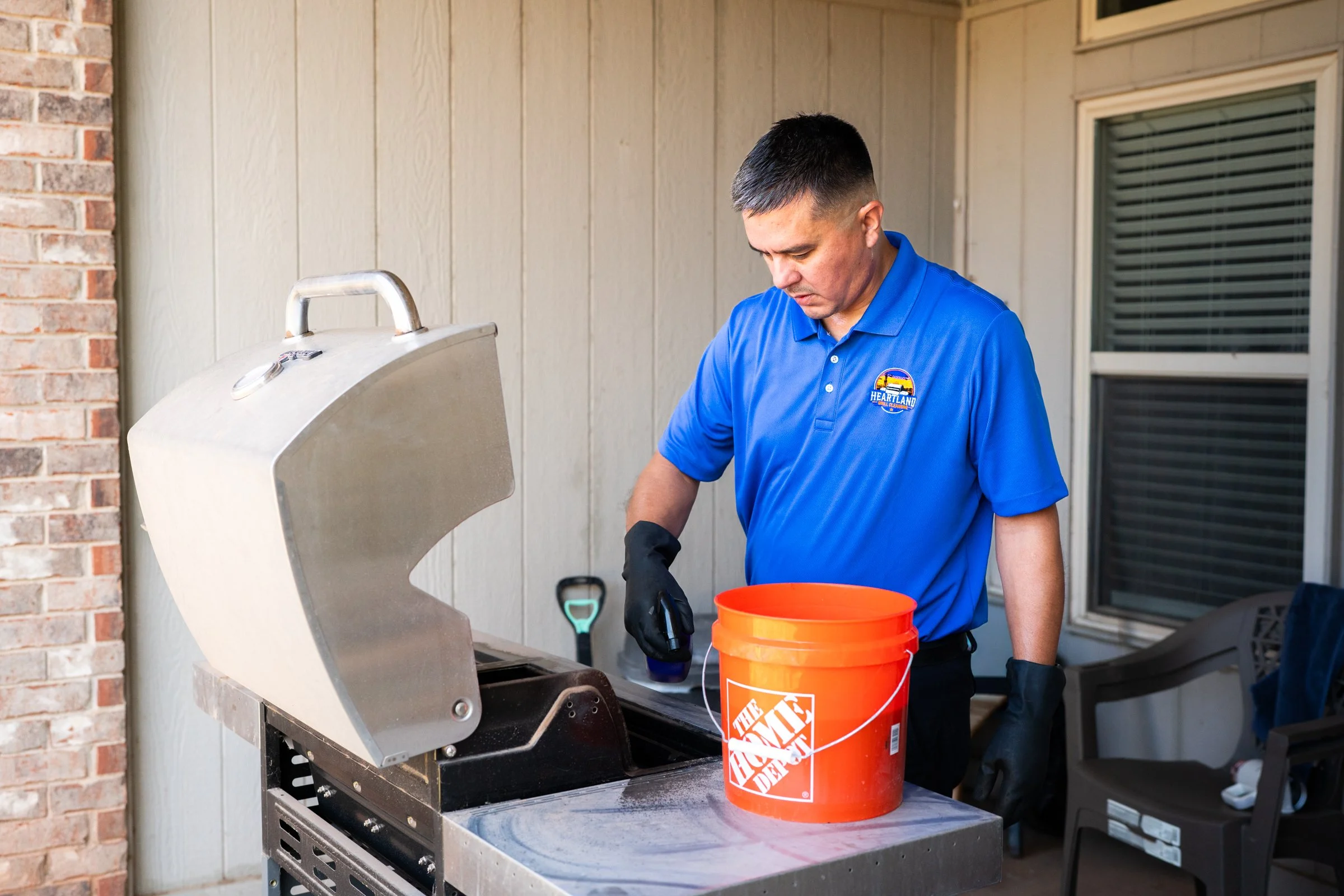A man in a blue uniform with a logo reading 'Heartland' on it is standing outdoors, grilling food on a smoker grill. He is looking down at the grill wearing black gloves, next to an orange bucket from The Home Depot.