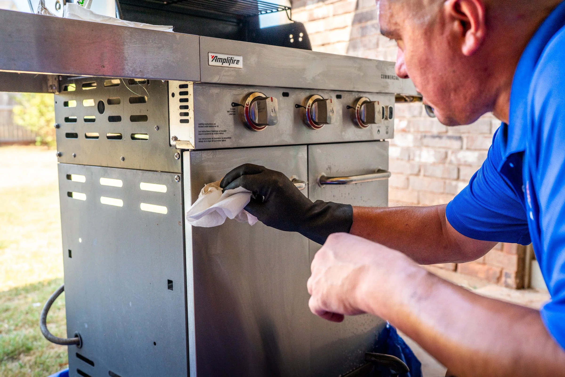 A person cleaning a large outdoor grill or stove using a cloth. The grill is made of metal and has control knobs on the front. The person is wearing a blue shirt and black gloves, with part of their face visible as they focus on cleaning the appliance. The background includes a brick wall and outdoor setting.