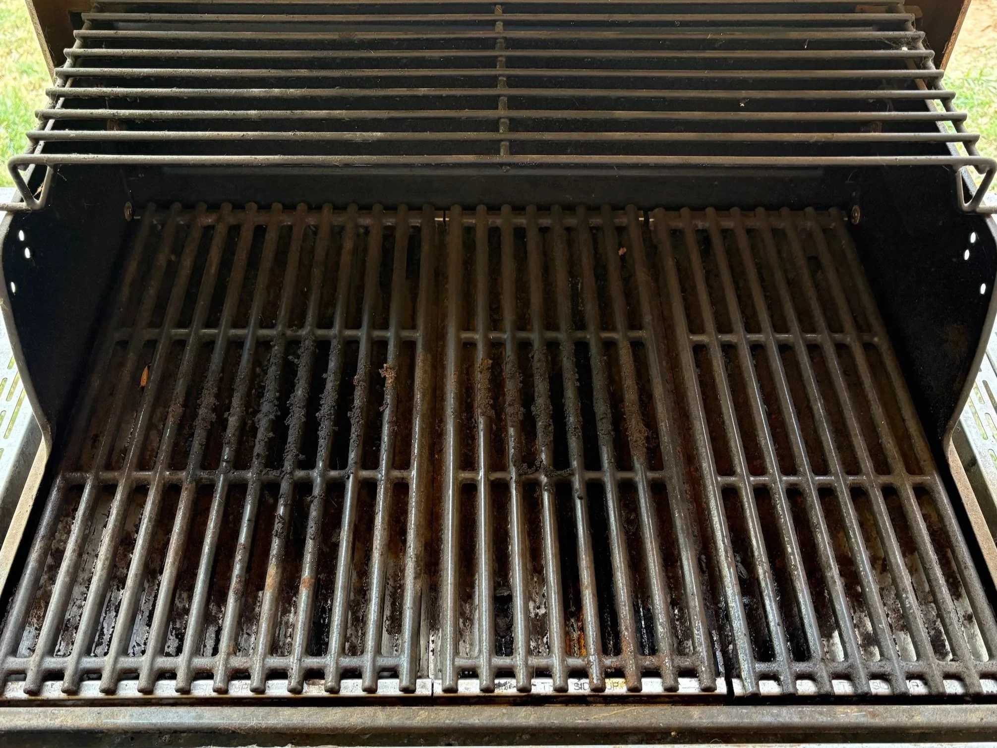Close-up of a black outdoor gas grill with a cooking grate, showing signs of grease and residue from previous use. The grill is outdoors with a hint of grass visible in the background.