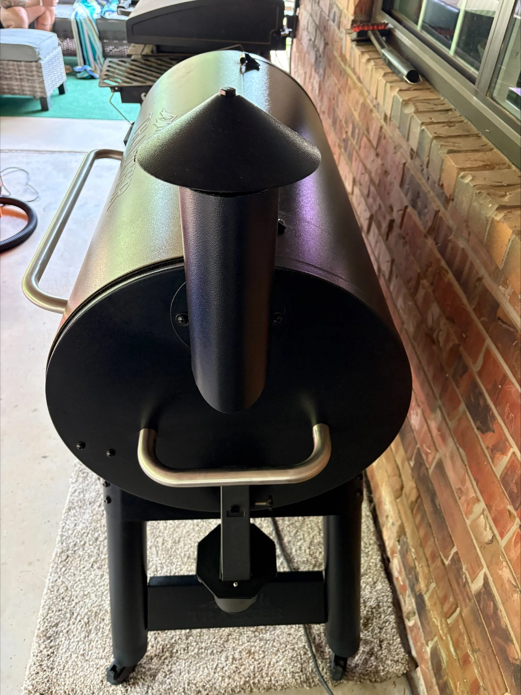 View of an outdoor black grill with a chimney, metal handles, and a brick wall behind it, positioned on a speckled white rug.