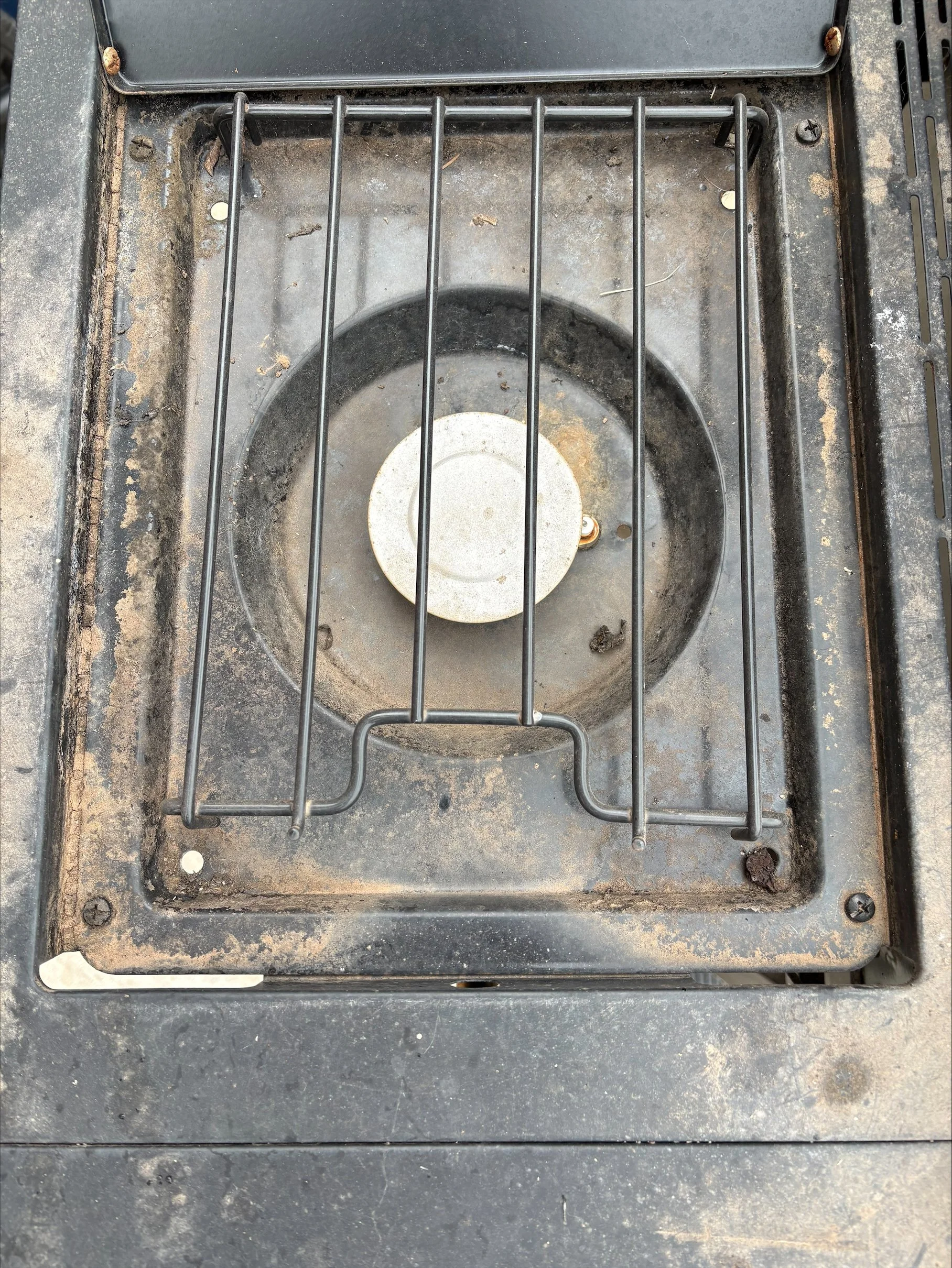 A close-up view of a dirty outdoor electric stove or cooktop with a metal grate over the circular burner area, showing signs of dirt, rust, and grime with some debris and small stains.