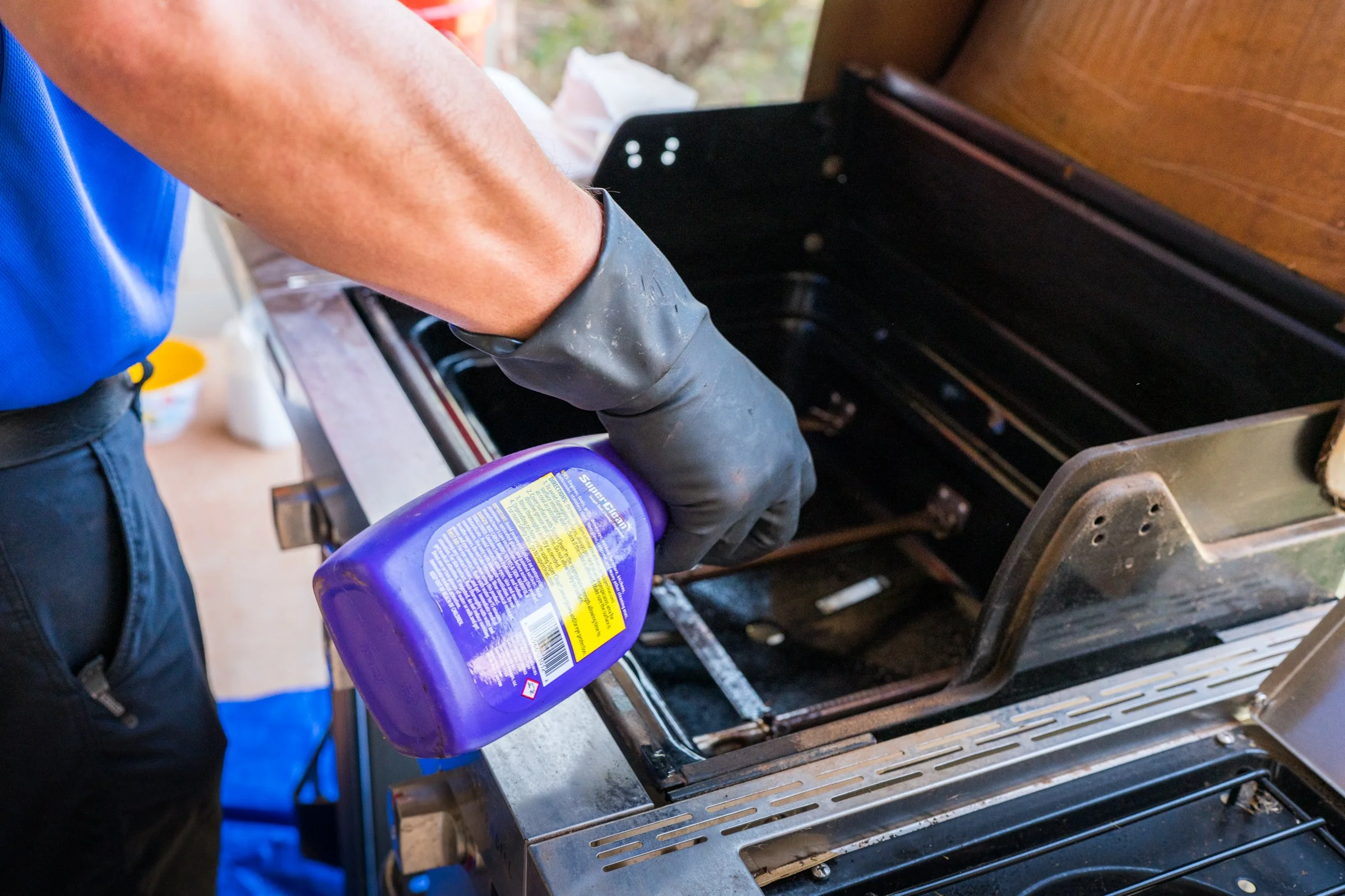 A person wearing a blue shirt and black gloves is holding a purple spray bottle and cleaning a black metal oven rack inside an open oven.