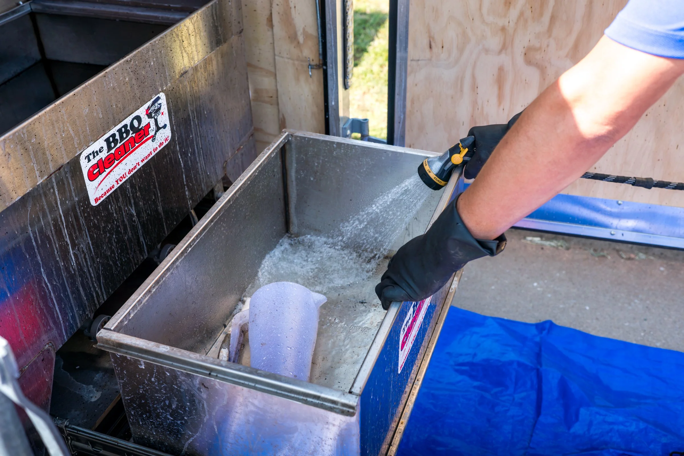 Person wearing a blue shirt and black gloves filling a metal cleaning sink with water attached to a dip-soaking container labeled 'The BBQ Cleaner'.
