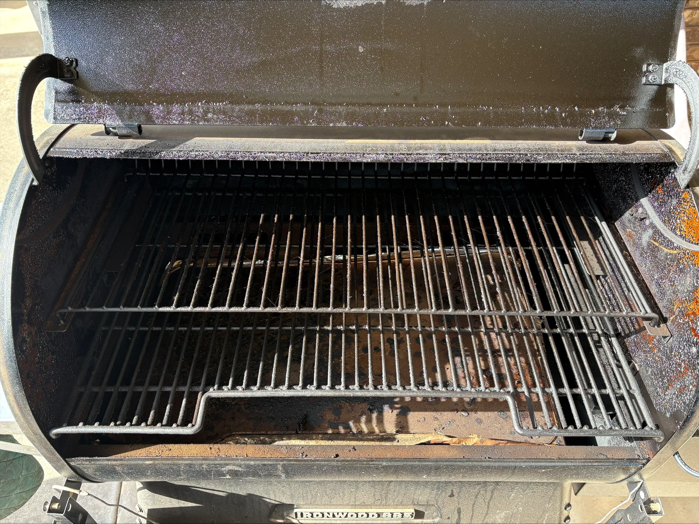 Empty grill with two cooking racks inside, some residue of ash and ash catchers visible, part of a wheel at the bottom left corner.