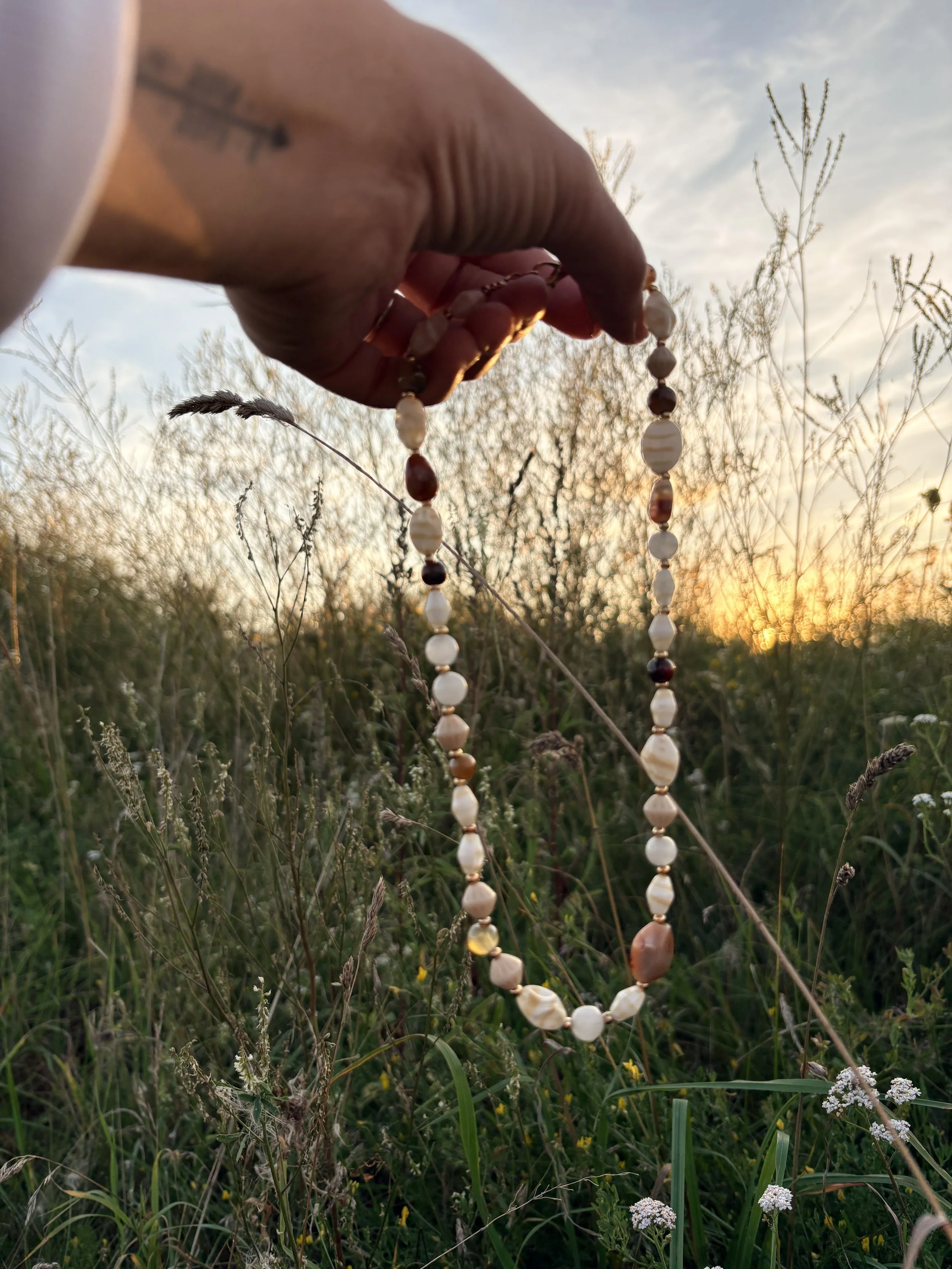 A person's hand holding a beaded necklace made of various shells and stones, with a field of wild grass and plants at sunset in the background.