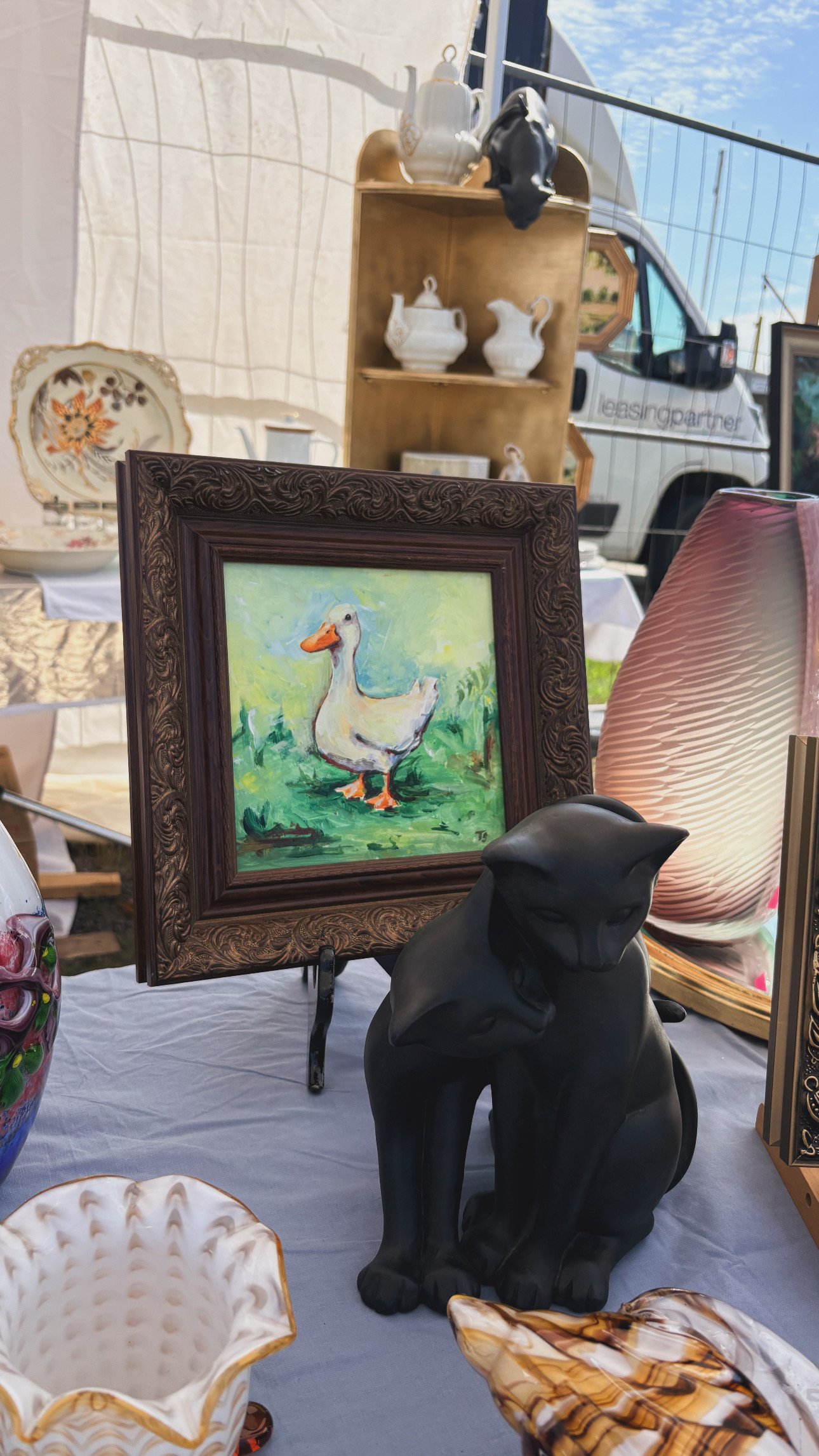 Various decorative items are displayed at an outdoor market, including a framed painting of a duck, a black sculpture of an animal, a large pink textured vase, and ceramic dishes with floral patterns. In the background, a wooden shelf holds white ceramic teapots and pitchers, and a vehicle with a partial logo is visible.