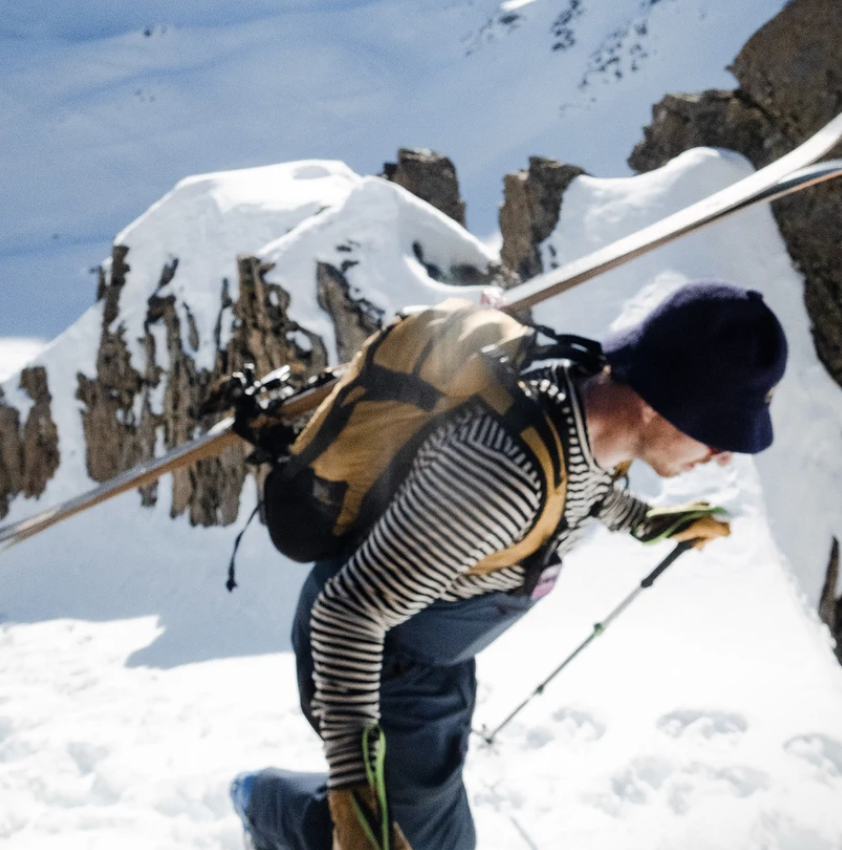Skier wearing a dark knit beanie carrying skis through snowy mountains