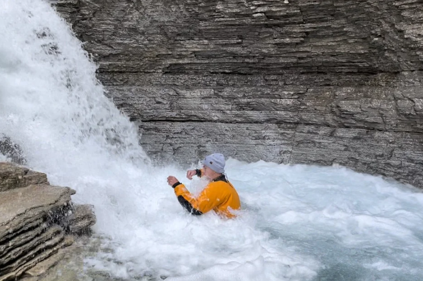 Person in an orange jacket sitting beside rushing mountain water and rock cliffs