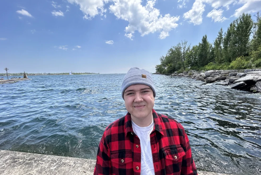 Person wearing a grey knit beanie and red plaid shirt beside a rocky lake