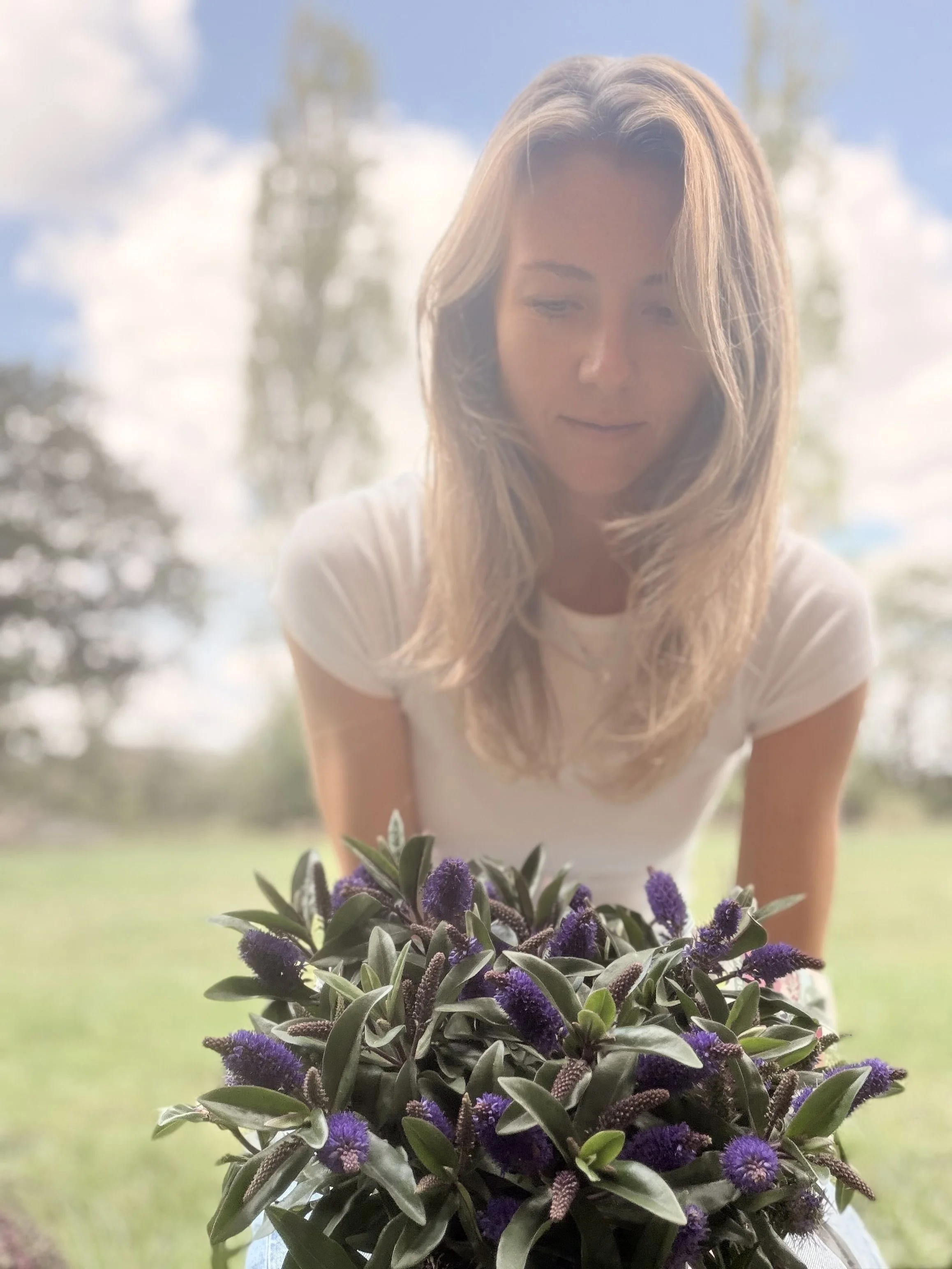A woman with long light brown hair looking down at purple flowers in her lap outdoors on a sunny day with trees and clouds in the background.