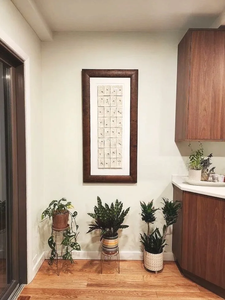 A cozy kitchen corner with three potted plants on wooden stands and floor, a framed artwork on the wall, and a wooden cabinet on the right.