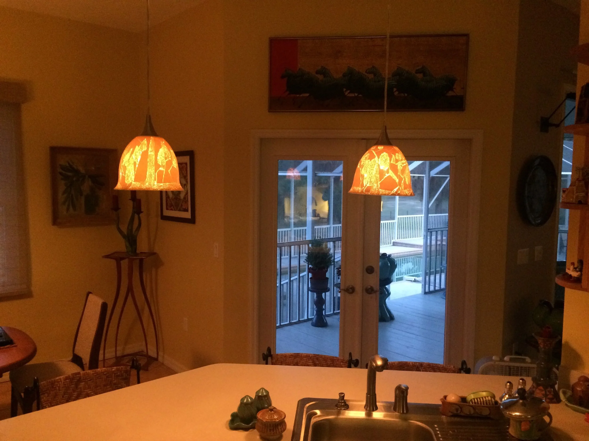 A cozy kitchen interior with orange pendant lights hanging over a counter, view through glass door to an outdoor balcony with potted plants, and artwork on the walls.