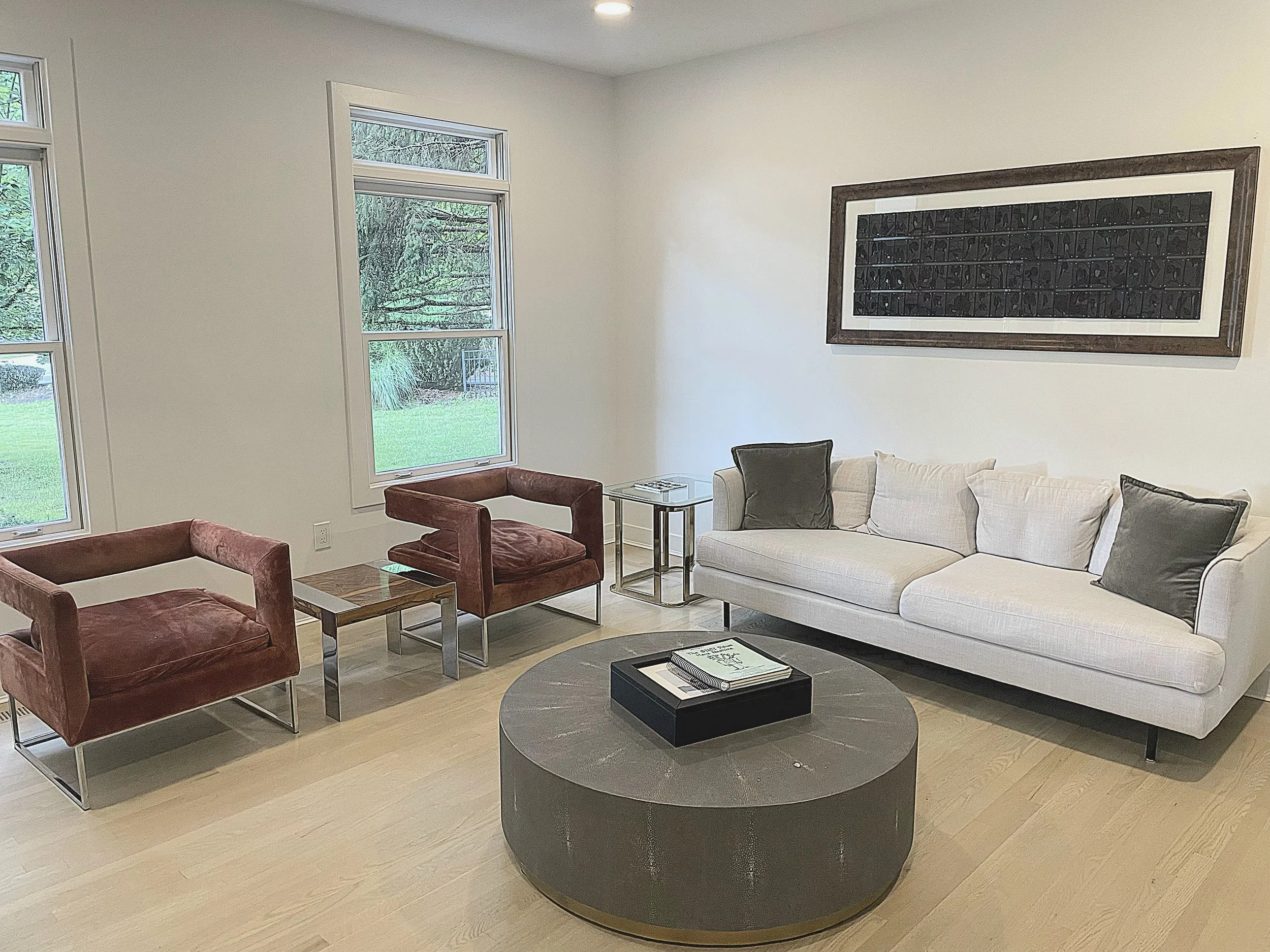 Living room with two large windows, a white sofa with dark pillows, two red armchairs, a round black coffee table with books, and a wall art piece with 88 carved black porcelain tiles in a walnut burl frame.