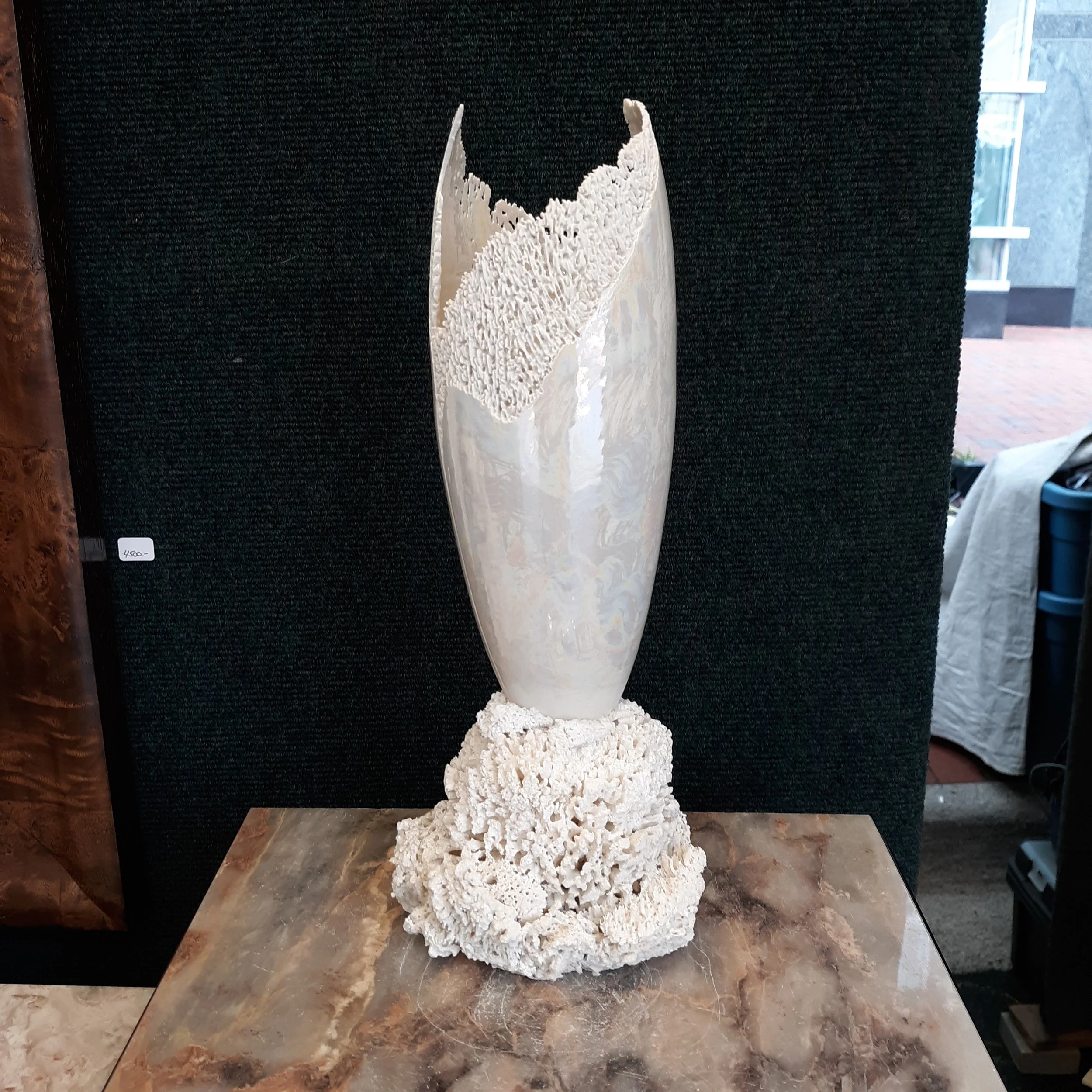 A tall, white coral sculpture with an intricate, porous surface stands on a base of similar coral at an art gallery. The background features a black textured wall and a large window letting in natural light.
