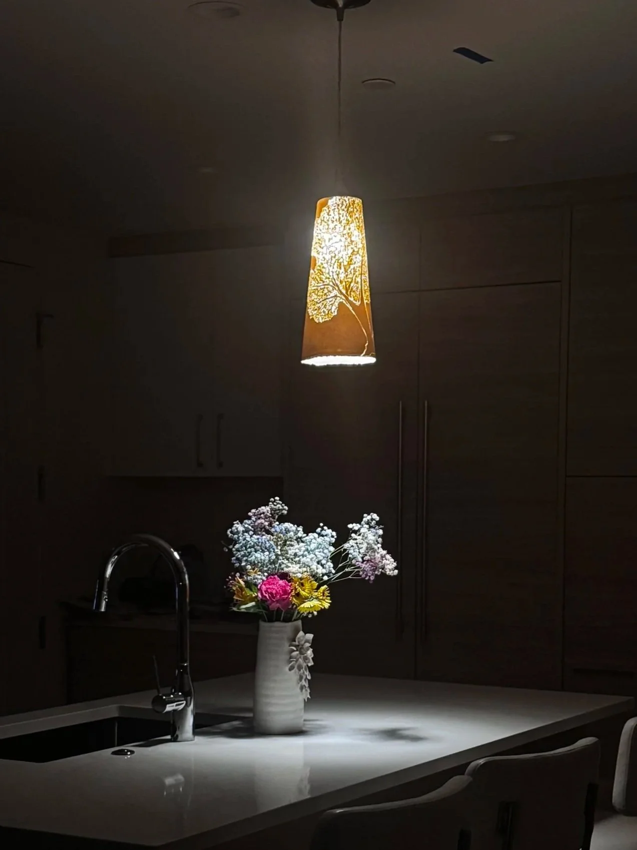 A dark room with a hanging light fixture featuring a tree design, a vase with assorted flowers on a kitchen counter, and kitchen cabinets in the background.