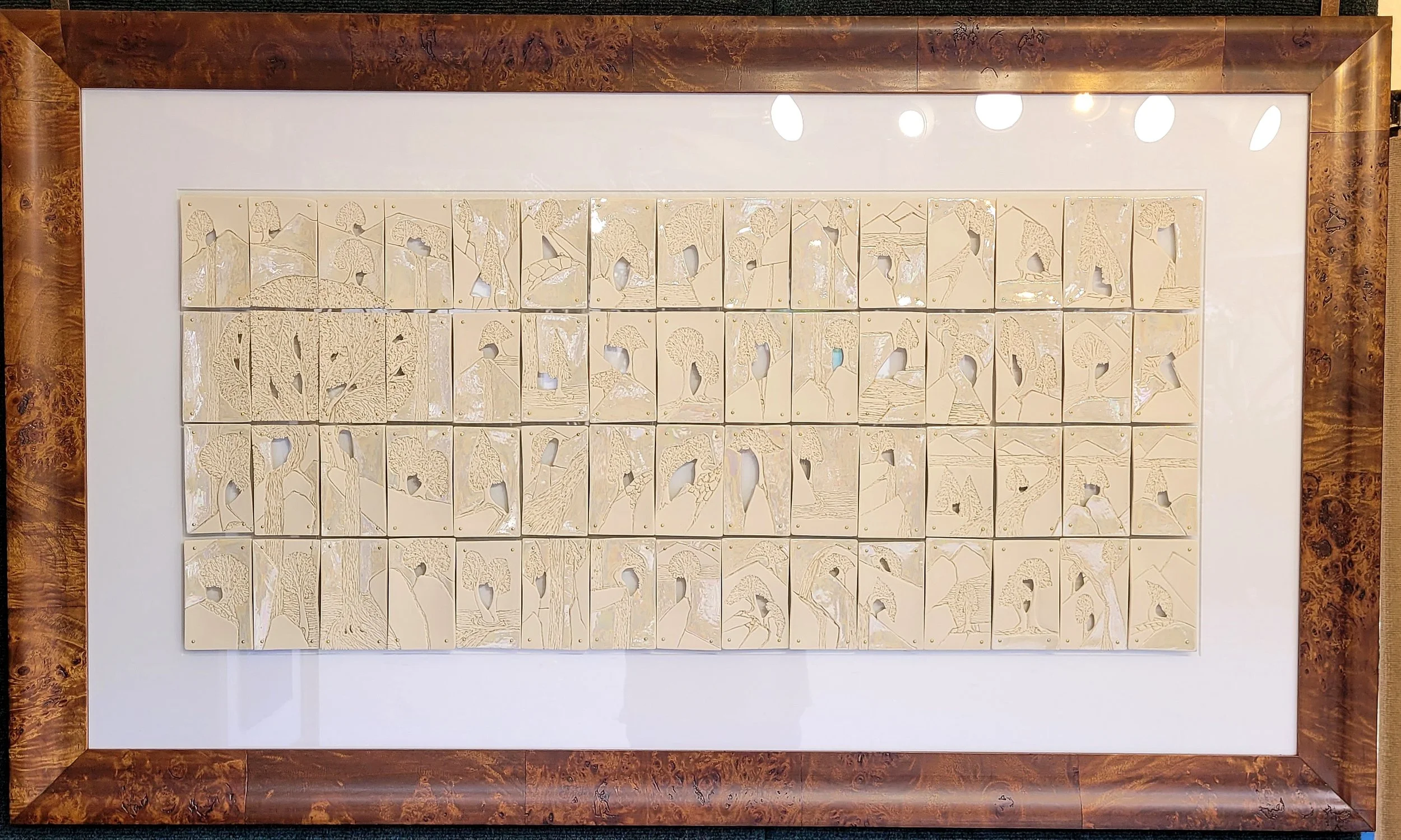 A framed wall art composed of multiple cream-colored tiles with engraved trees and mountain landscape designs, mounted on a white background in a chestnut burl frame.