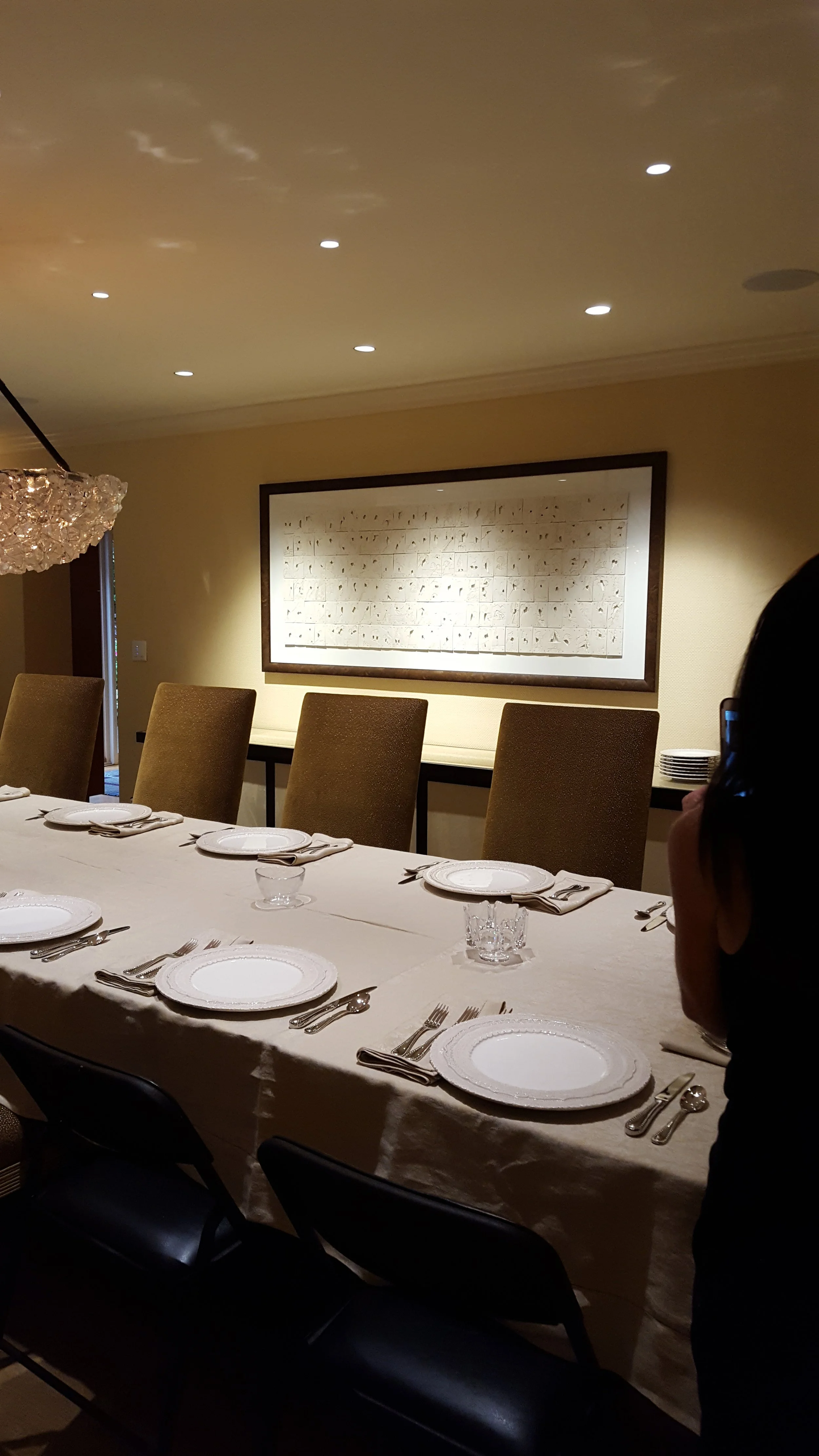 A dining room with a long table set with plates, napkins, utensils, and glasses, surrounded by green chairs, with a framed art piece on the wall and ceiling lights.
