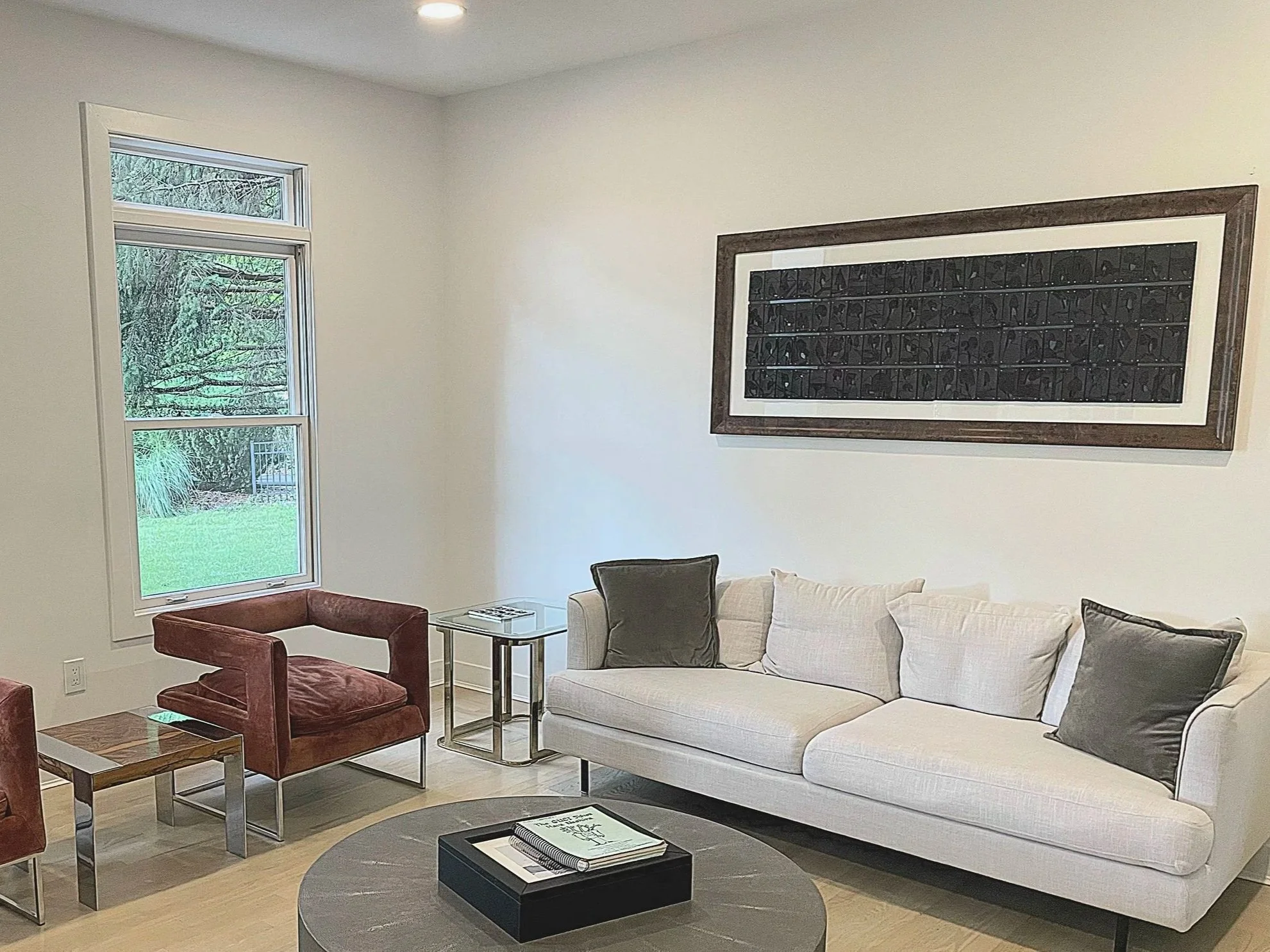A modern living room featuring a large white sofa with pillows, a round gray coffee table with books on it, a vintage-style armchair, a small side table, and a framed black art piece on the wall. There is a window showing greenery outside.