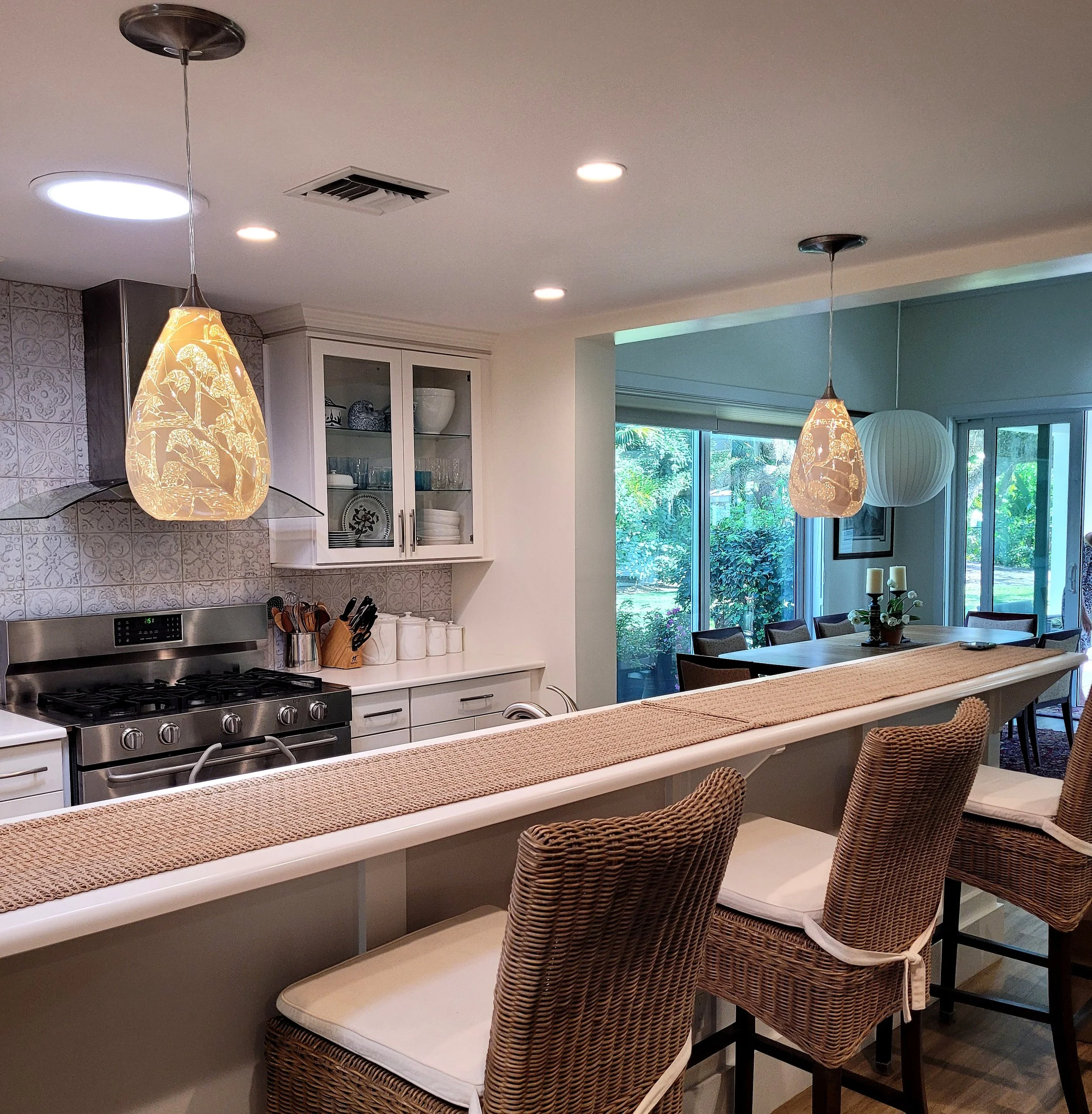A kitchen with hanging pendant lights over a white countertop island, a stainless steel stove, glass-front cabinets, and a dining area with chairs and a large window overlooking greenery outside.