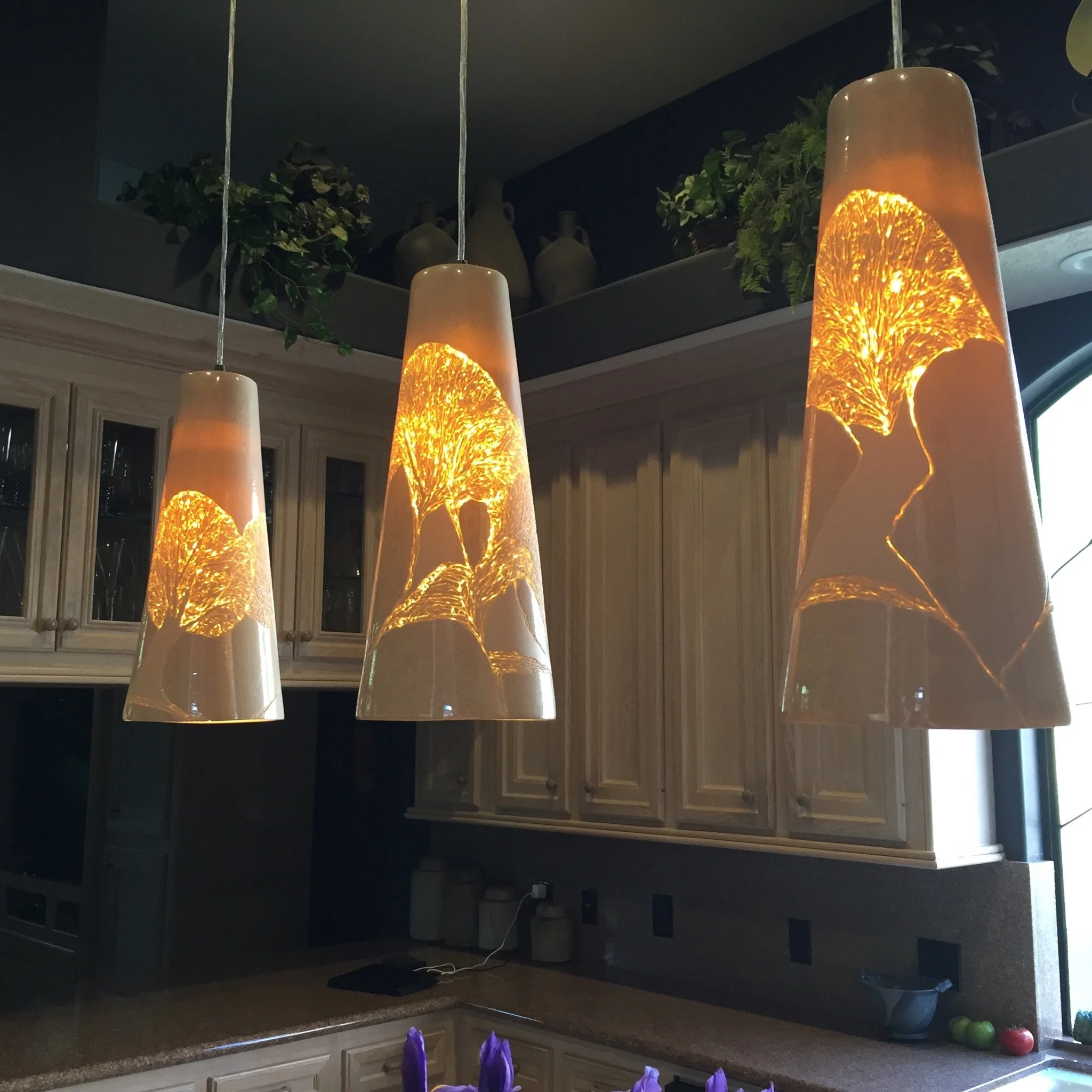 Kitchen with illuminated pendant lights shaped like slices of citrus fruit hanging above a dark countertop. The upper part of the kitchen has cabinets with glass doors and decorative vases and greenery on the shelves. A portion of the window and some apples on the counter are also visible.