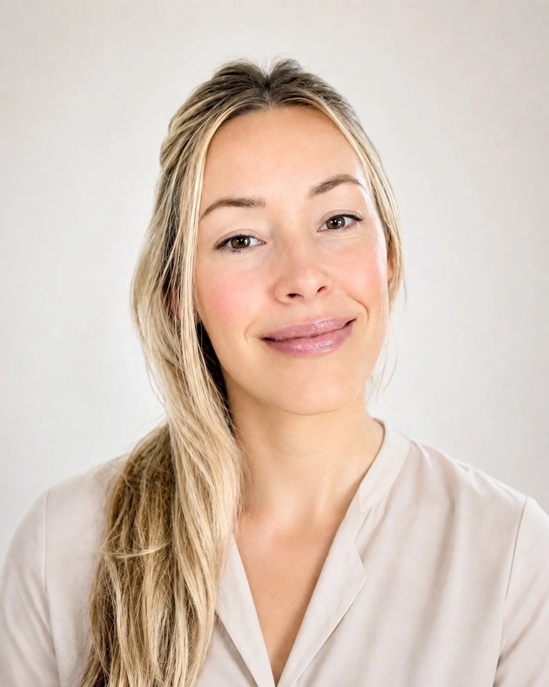 A woman with long blonde hair and light skin smiling softly, wearing a light-colored shirt against a plain background.